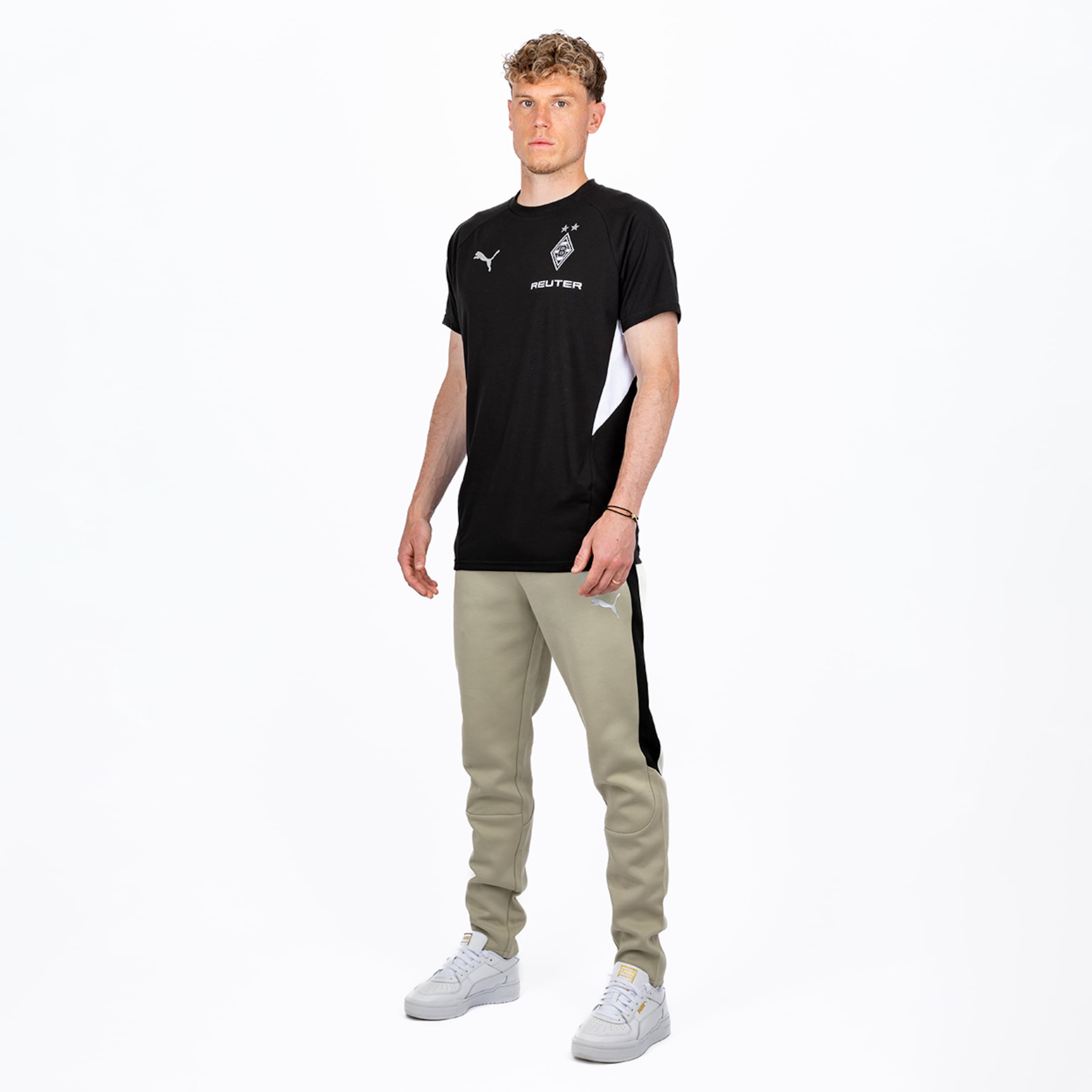 Ein junger Mann mit Locken trägt ein schwarzes Puma T-Shirt mit Borussia Mönchengladbach Logo und 'REUTER'-Sponsor, hellolivfarbene Trainingshose und weiße Sneaker vor weißem Hintergrund.