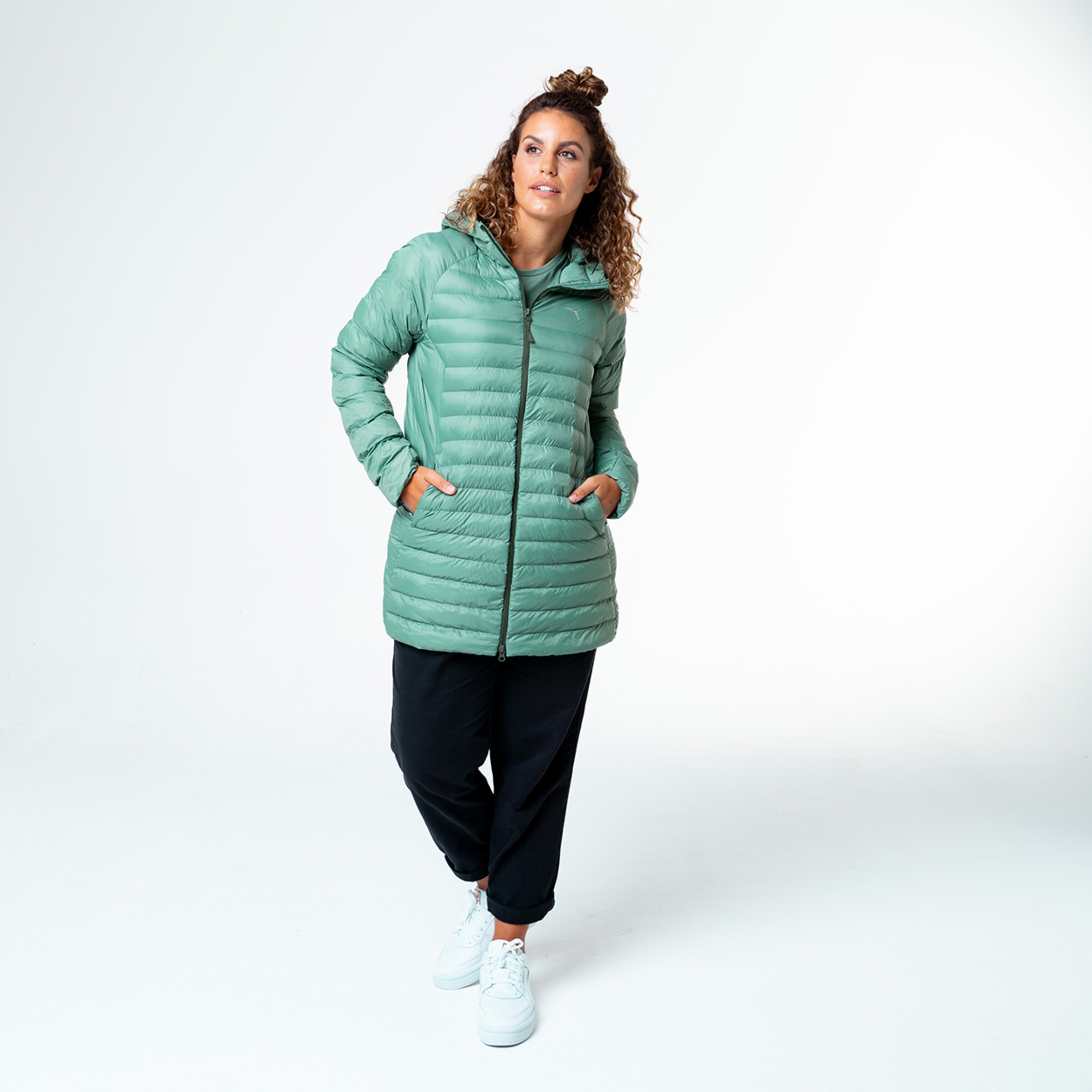 Frau trägt eine grüne Pufferjacke und schwarze Hose lächelnd und stehend.