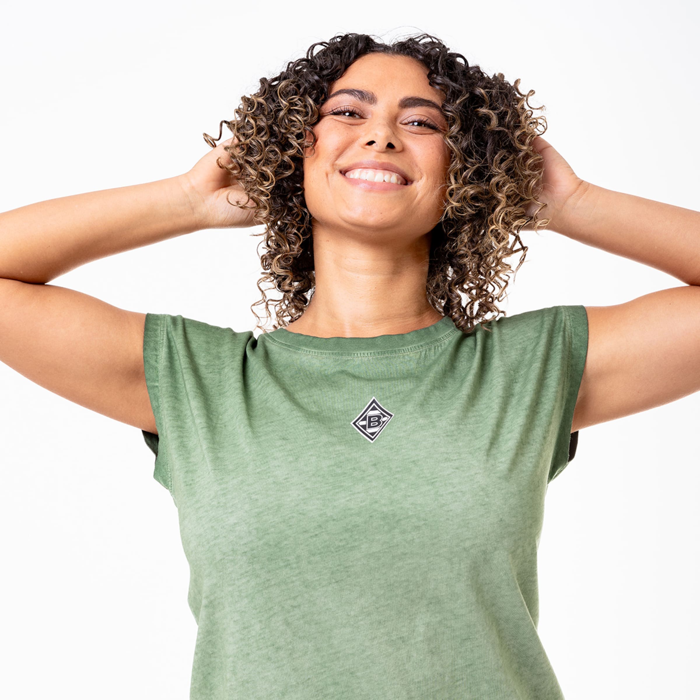 Frau mit lockigem Haar lächelt und hält ihre Hände hinter dem Kopf trägt ein grünes T-Shirt mit Vereinslogo.