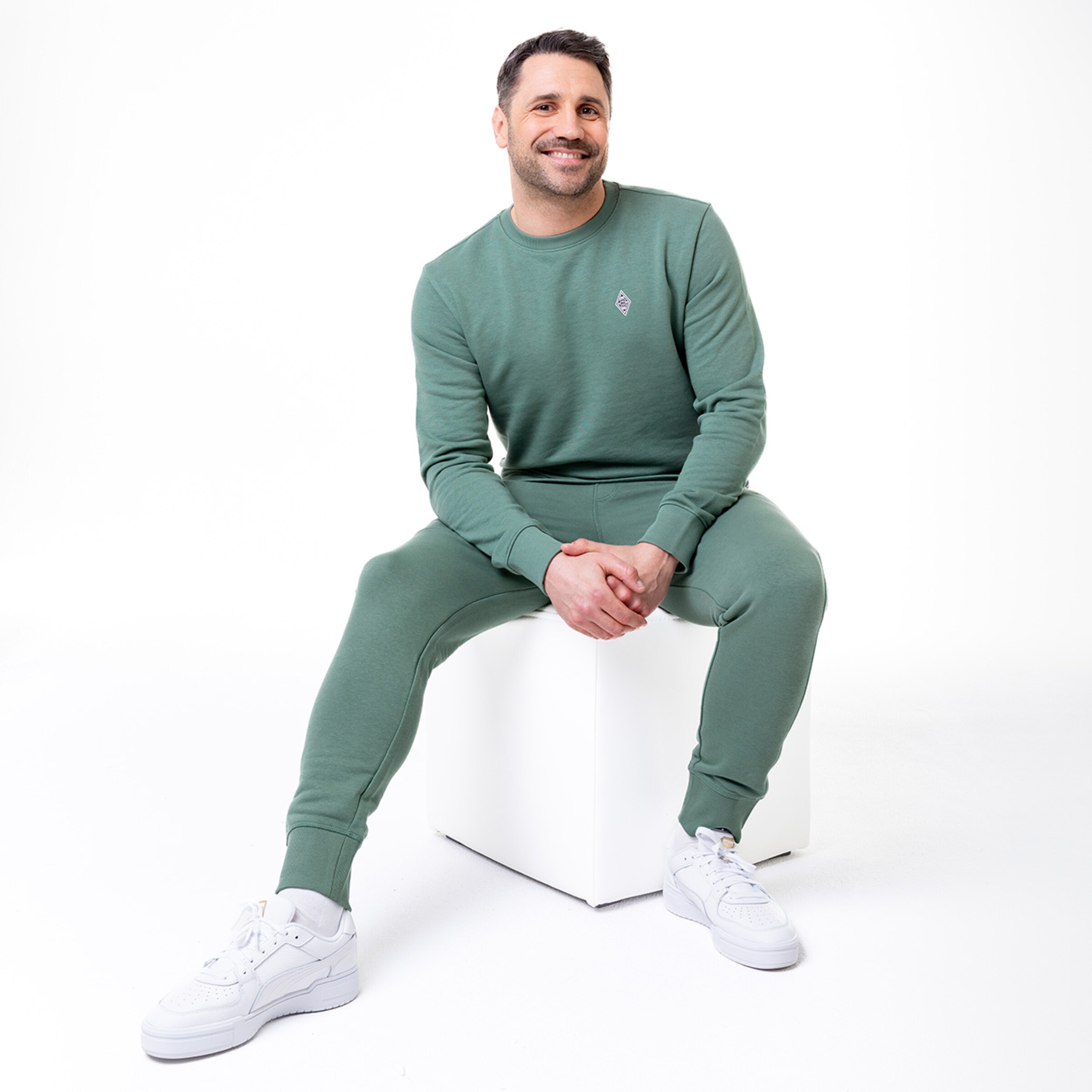 Mann in grauem Trainingsanzug sitzt auf einem weißen Würfel und lächelt.