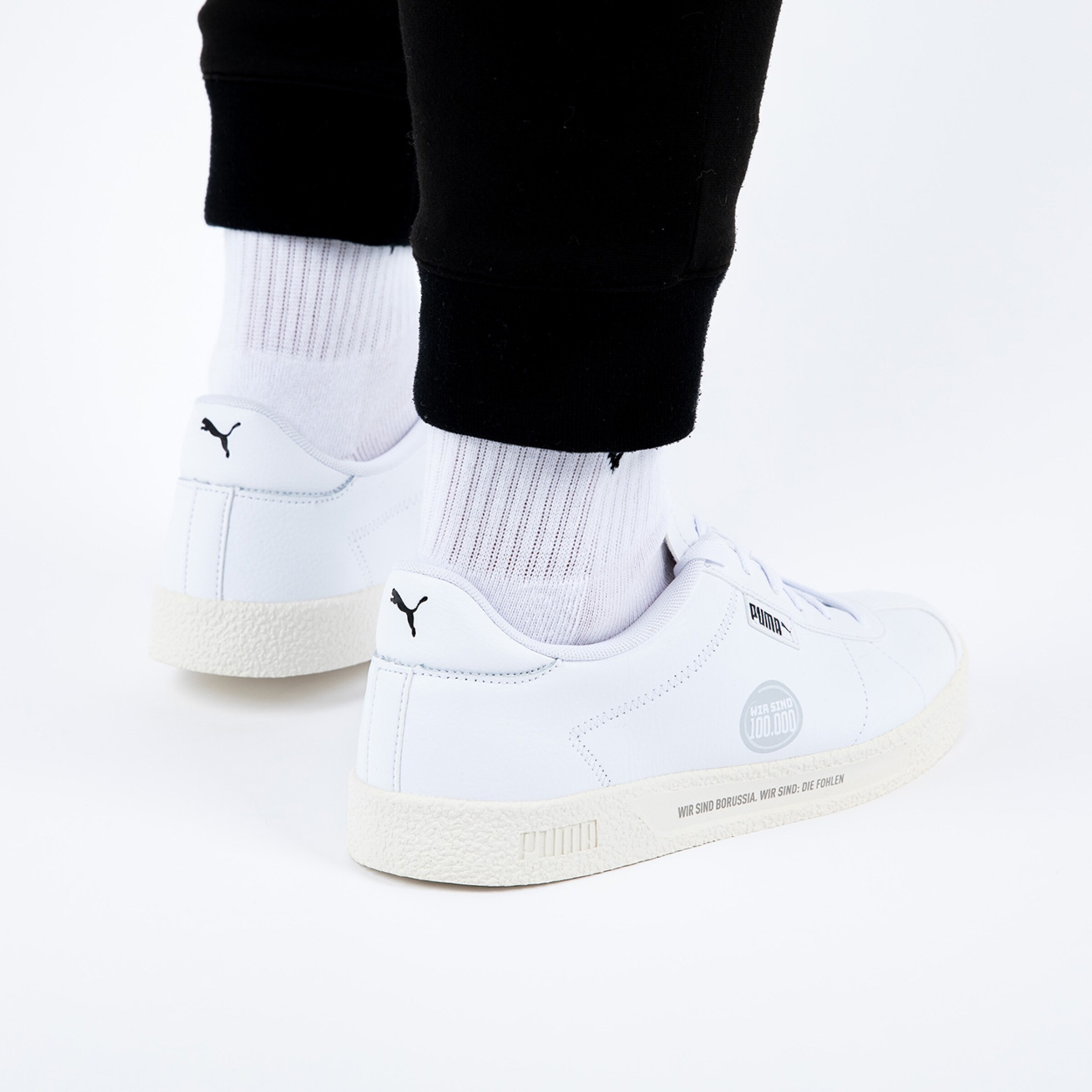 Weiße Puma-Sneaker mit grauem Logo getragen mit schwarzen Hosen und weißen Socken.