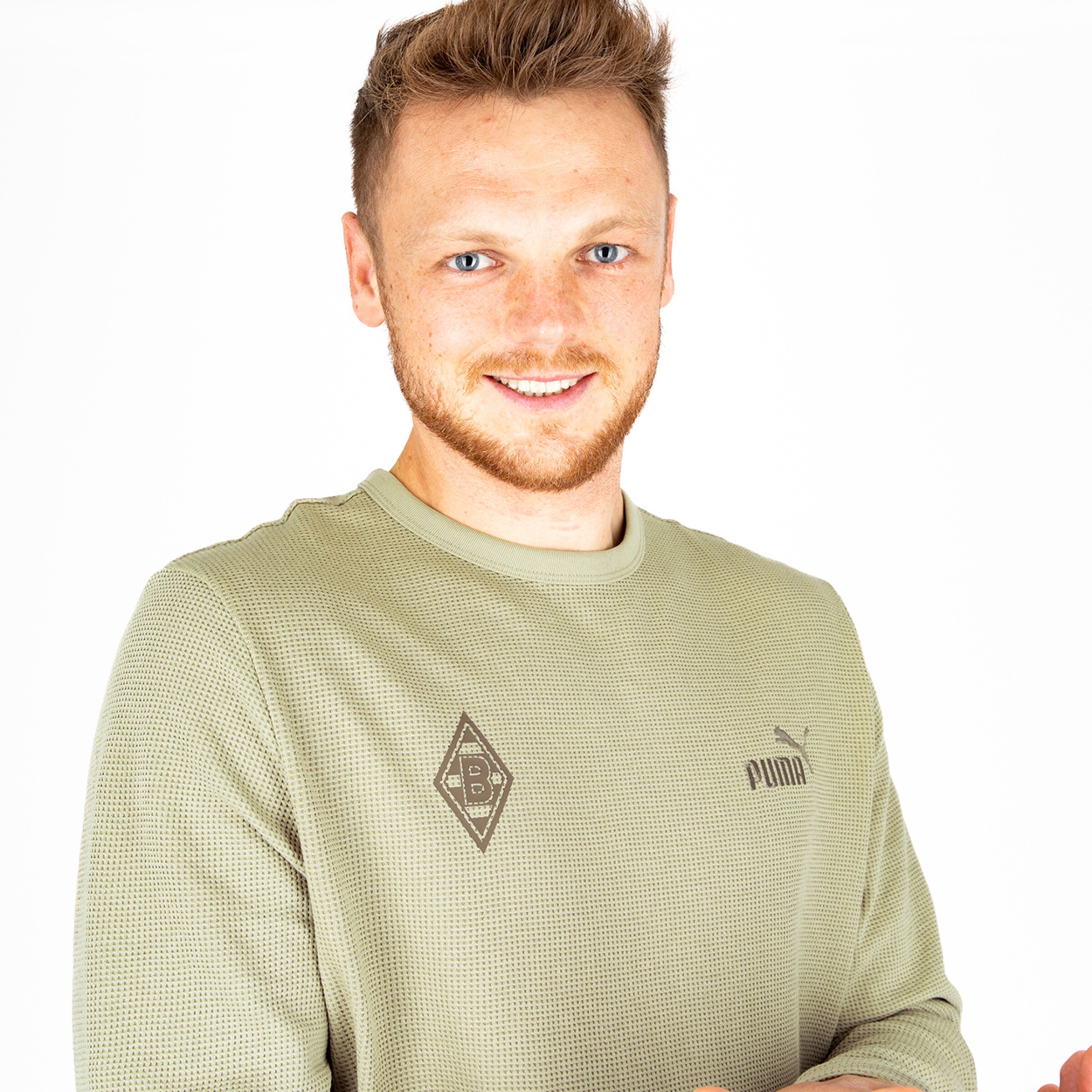 Junger Mann mit blondem Haar der ein graues Puma-Oberteil mit Borussia Mönchengladbach-Logo trägt und lächelt.