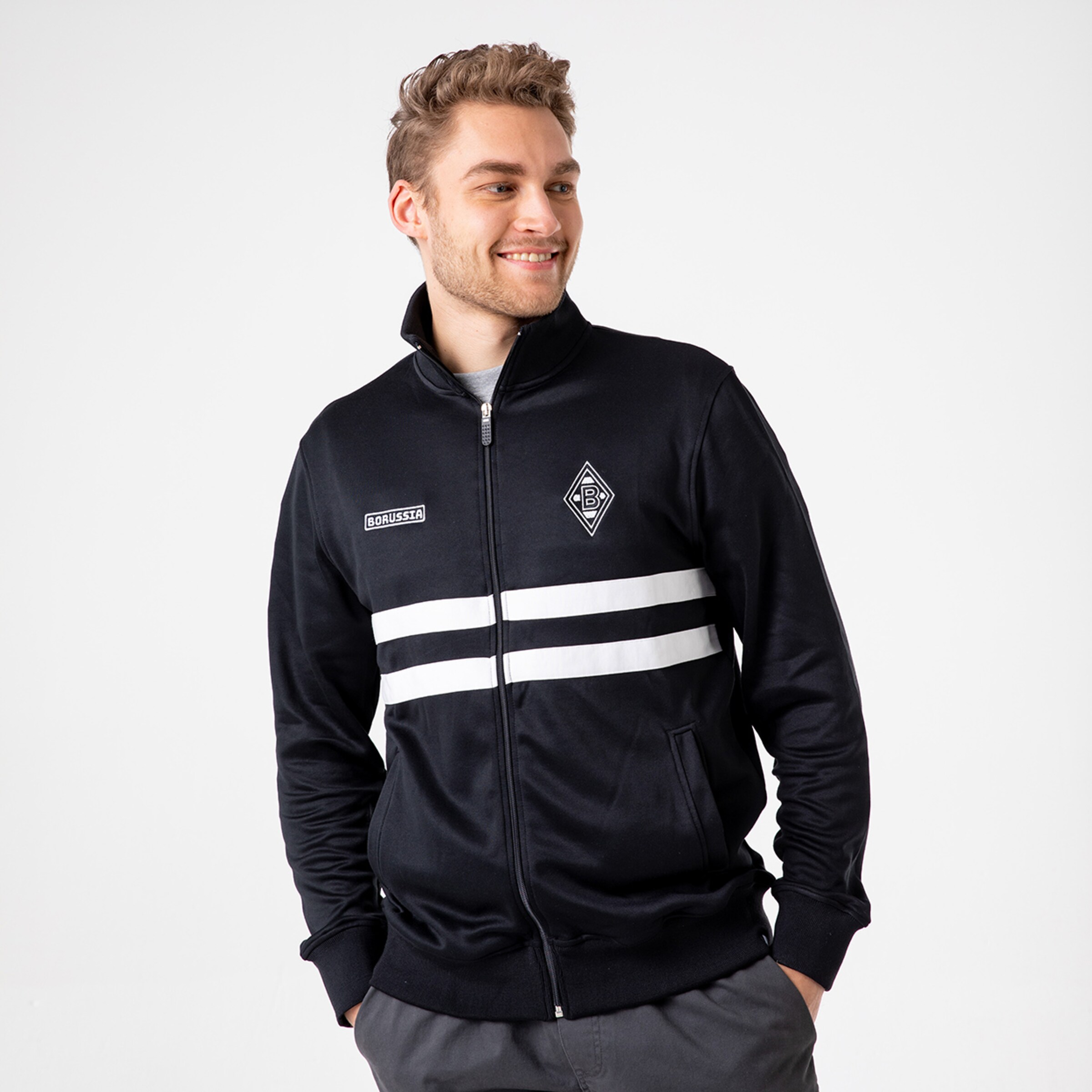 Ein Mann mit einem schwarzen Trainingsanzug mit weißen Streifen und Borussia Mönchengladbach-Logo steht lächelnd.