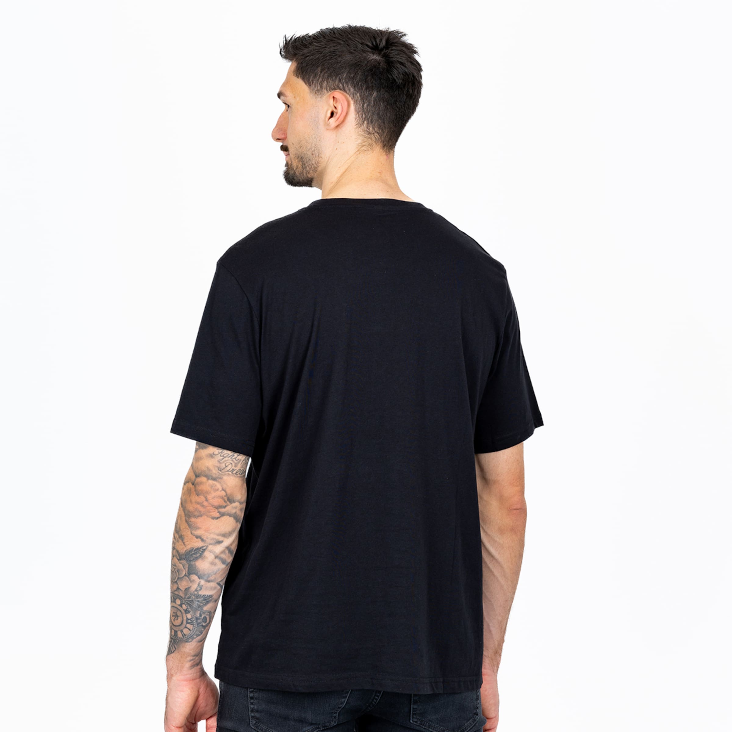 Rückansicht eines Mannes in schwarzem T-Shirt vor weißem Hintergrund. Sichtbare Tätowierung am linken Arm.