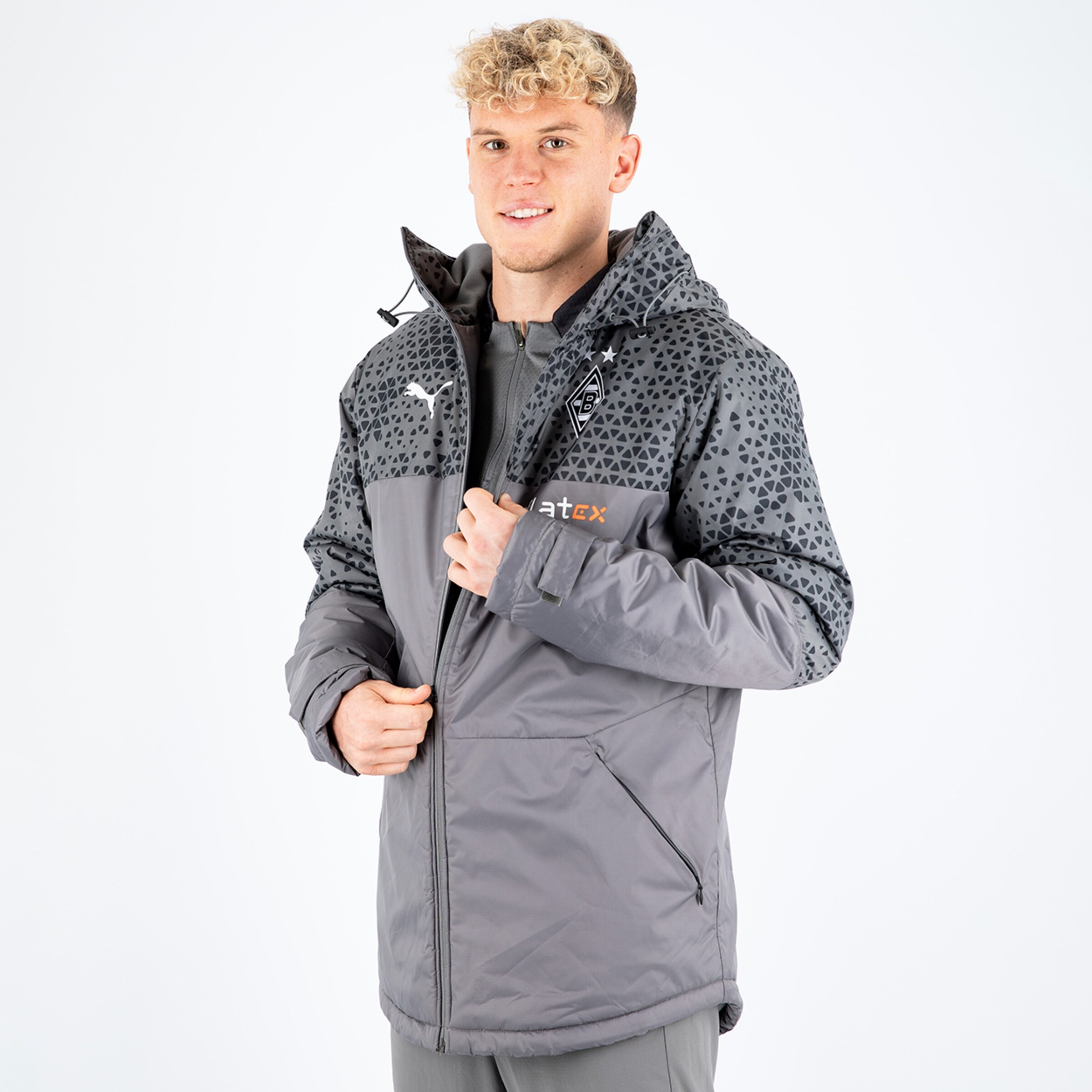 Ein junger Mann trägt eine graue Jacke mit Muster und steht lächelnd in Pose.