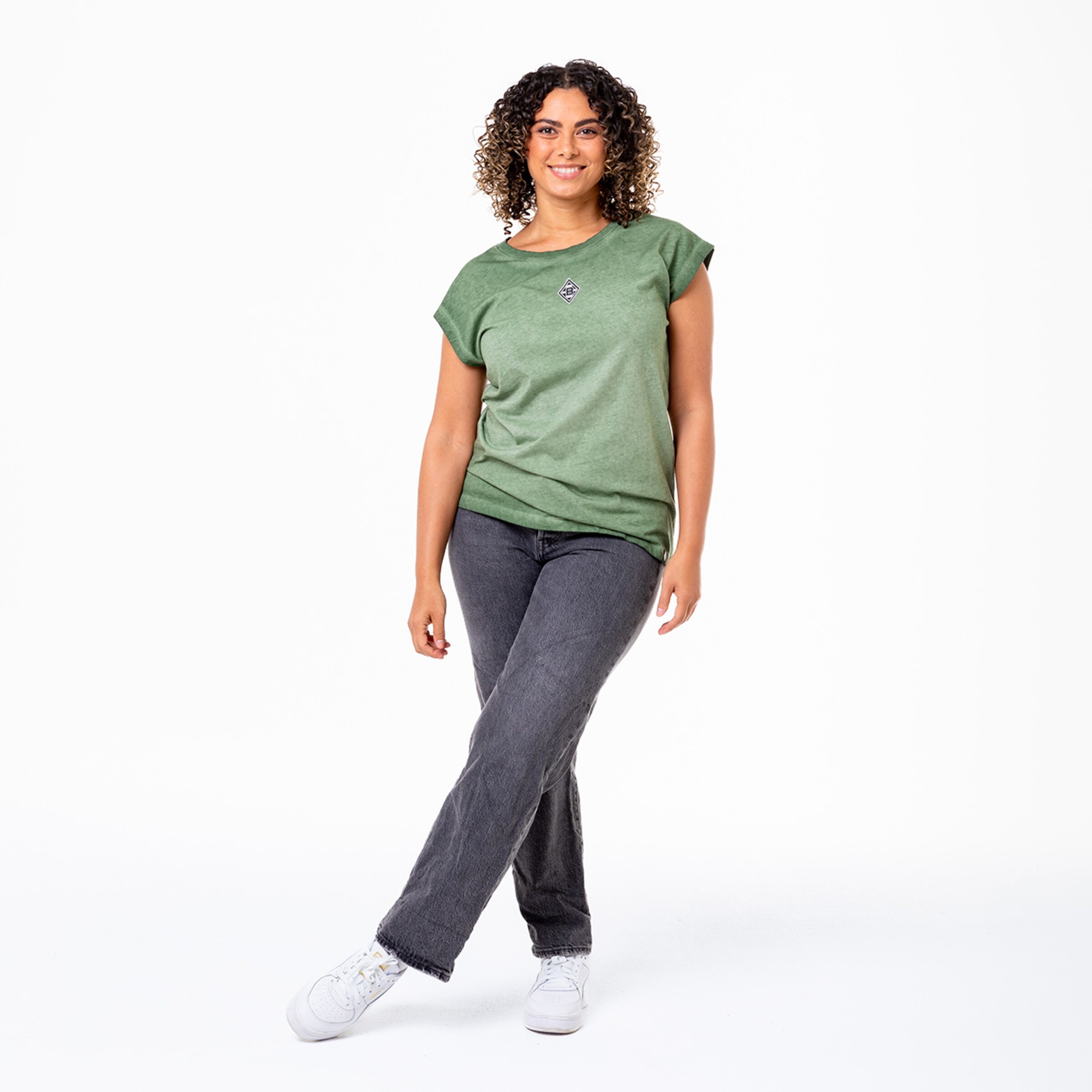 Frau mit lockigem Haar trägt ein grünes T-Shirt und graue Jeans.