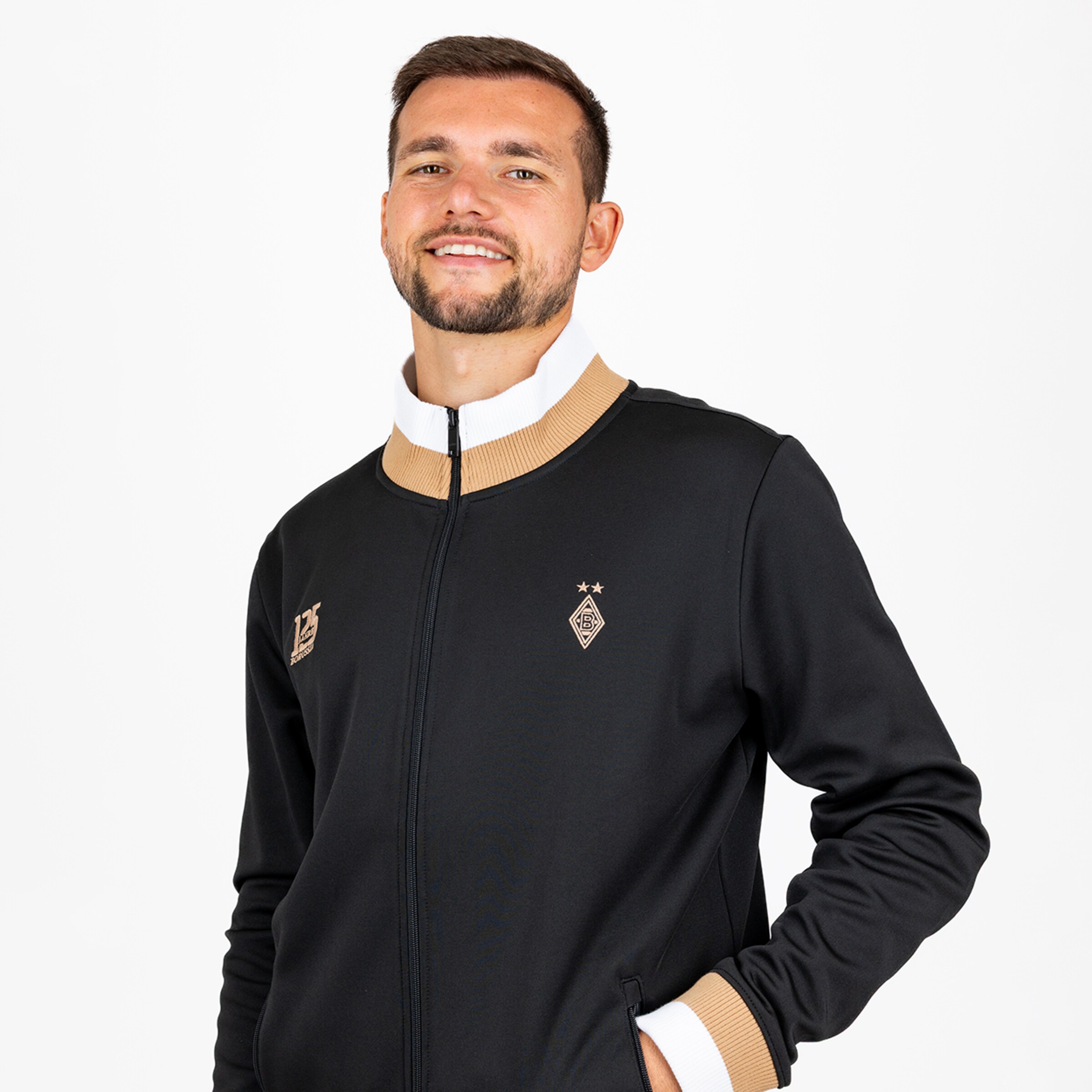 Lächelnder Mann in schwarzer Borussia Mönchengladbach Trainingsjacke mit gold-weißem Kragen und 125-Jahre-Jubiläumslogo.