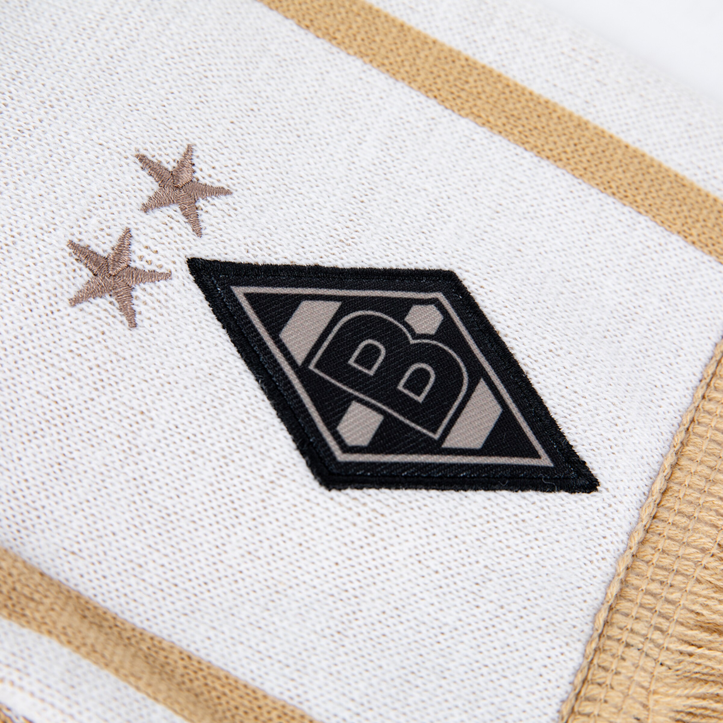Nahaufnahme des gestickten Borussia Mönchengladbach Logos mit zwei Sternen auf weißem Strickgewebe mit goldenen Akzenten.