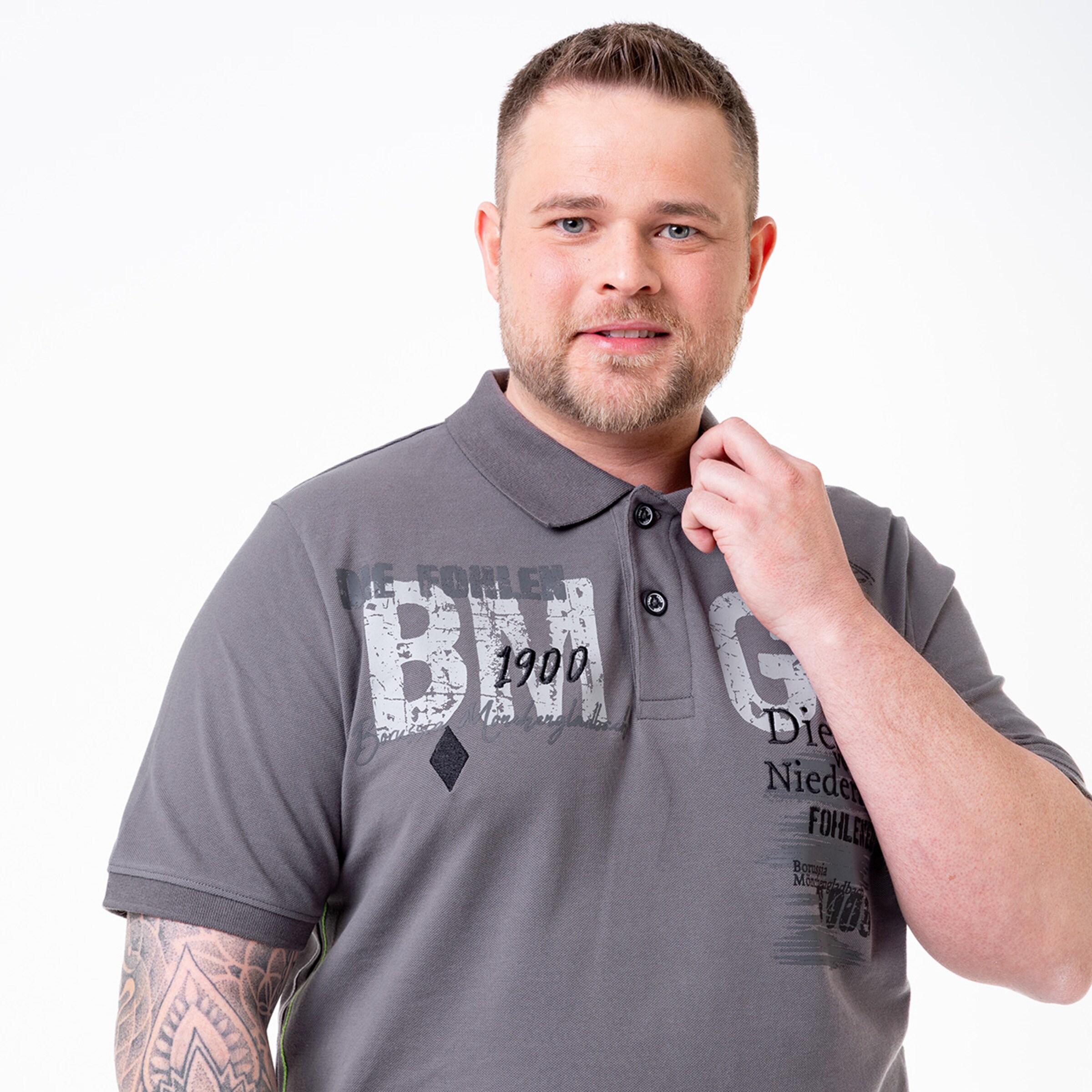 Mann mit tätowiertem Arm trägt ein graues Poloshirt zeigt ein freundliches Lächeln und greift an seinen Hals.