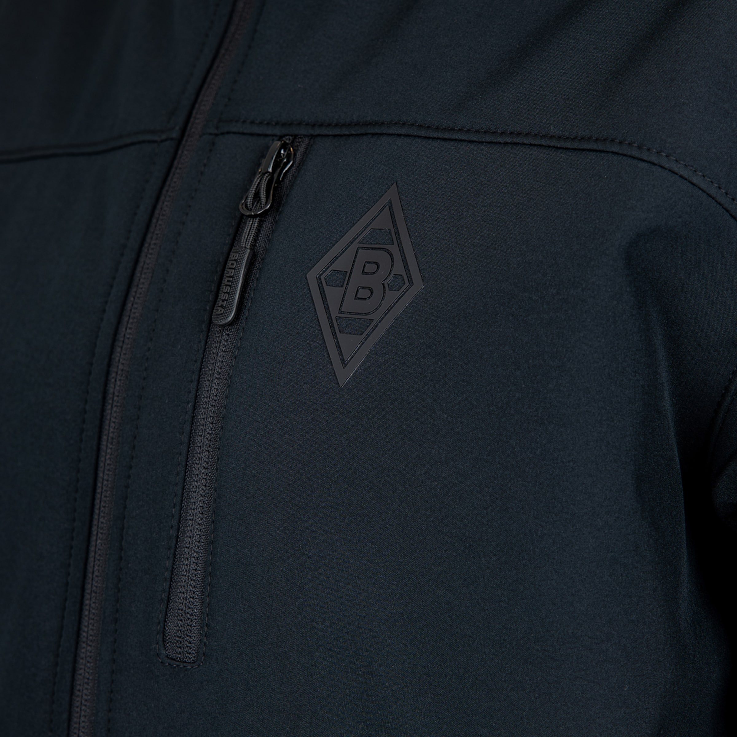 Nahaufnahme einer schwarzen Jacke mit Borussia Mönchengladbach-Logo (Raute) und einer vertikalen Brusttasche mit Reißverschluss.