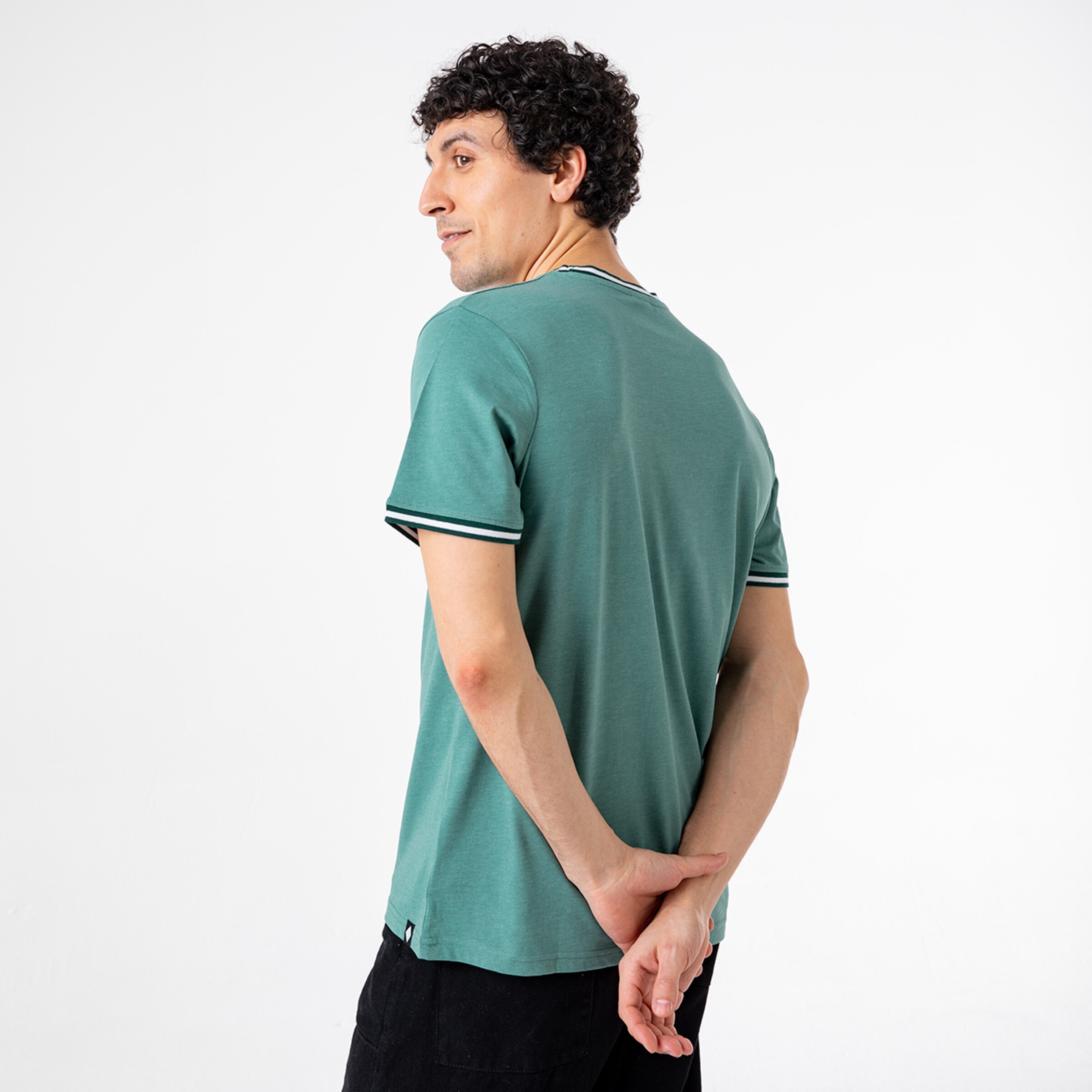 Man with curly hair wearing a teal t-shirt with striped sleeves stands with his back to the camera hands clasped behind his back.