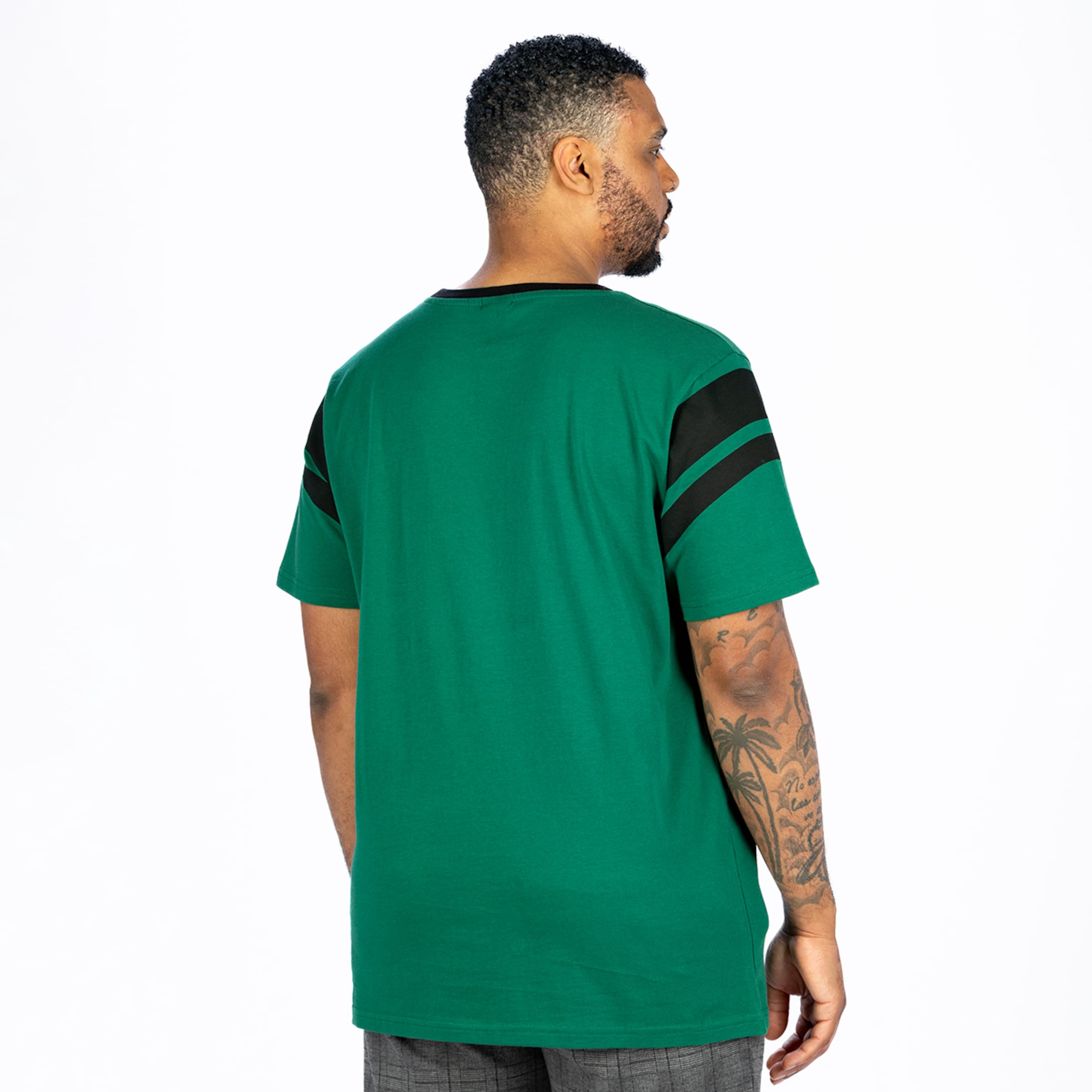 Rückansicht eines Mannes in einem grünen T-Shirt mit schwarzen Ärmelstreifen, der nach rechts blickt. Er hat einen Bart und Armtattoos mit Palmen und Text. Weißer Hintergrund.
