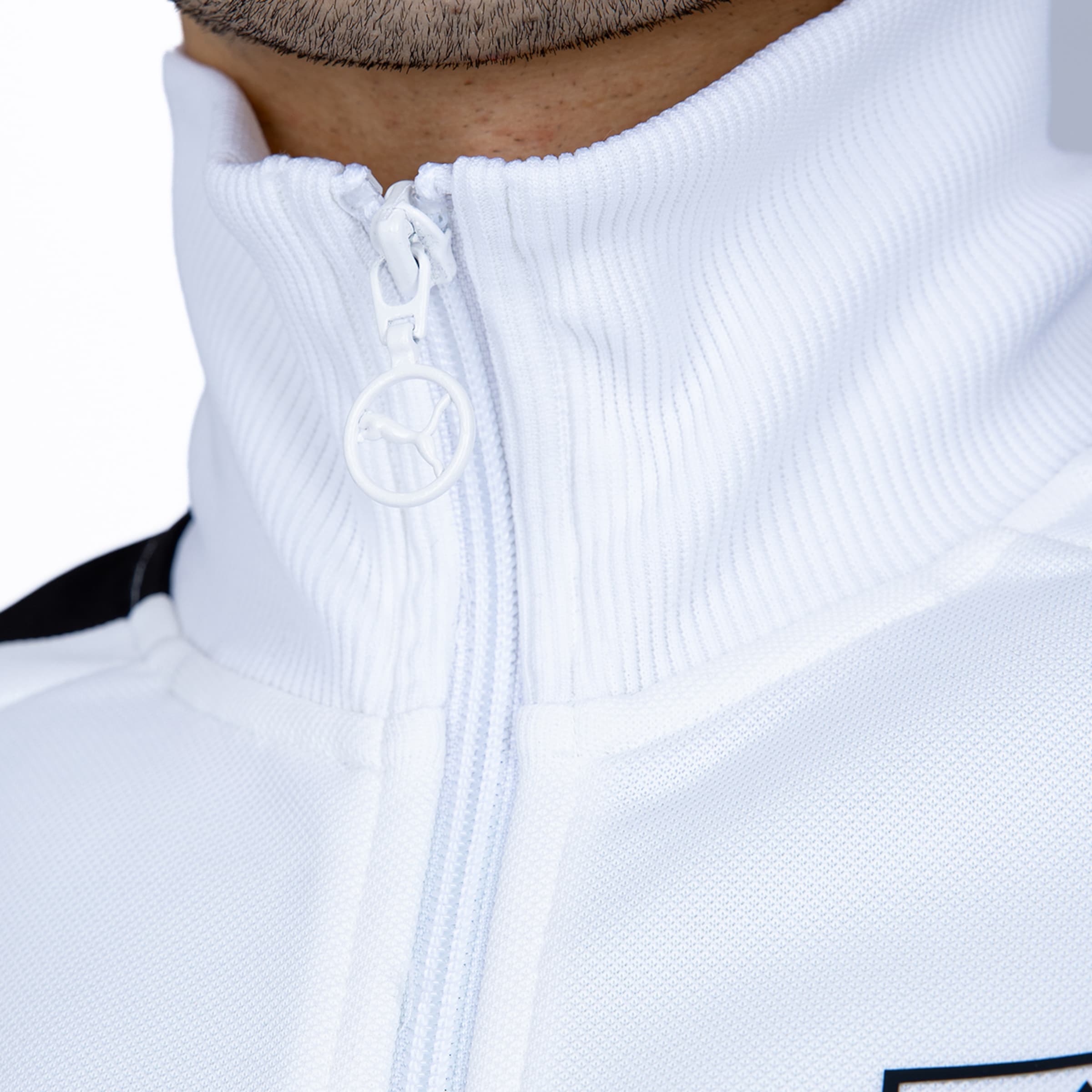 Nahaufnahme einer weißen Trainingsjacke mit geripptem Stehkragen und kreisförmigem Puma-Logo am Reißverschluss.
