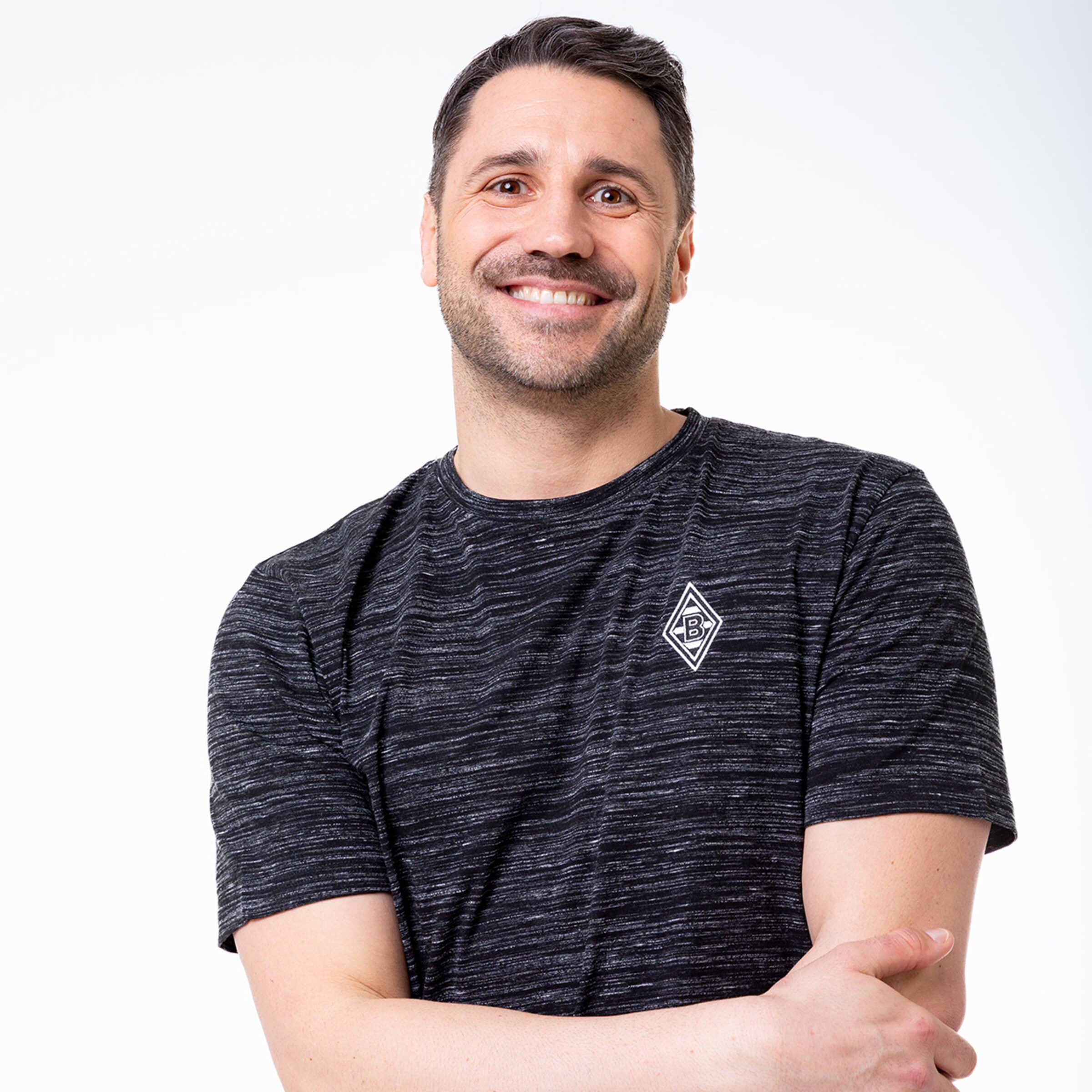 Ein Mann mit mittellangen dunklen Haaren lächelt freundlich und trägt ein schwarzes gestreiftes T-Shirt mit dem Borussia Mönchengladbach-Logo auf der linken Brust.