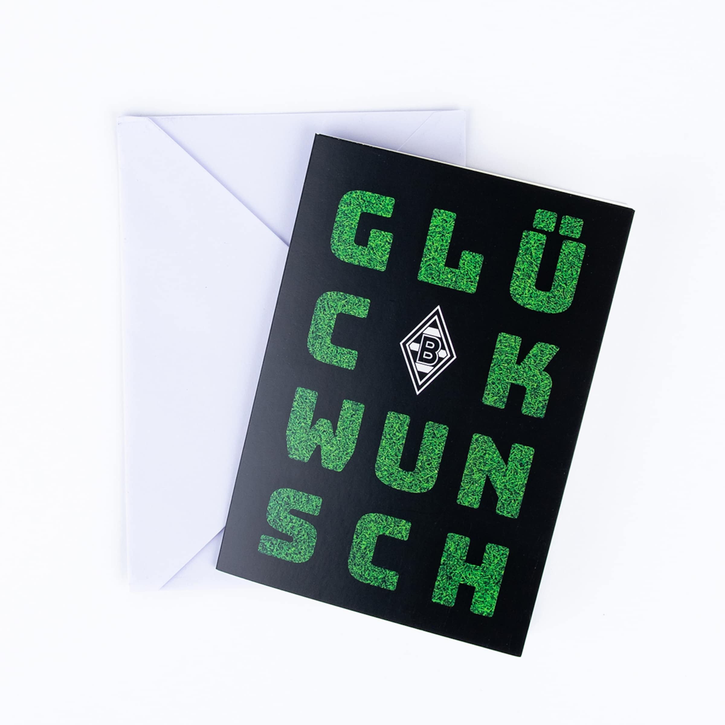 A black greeting card featuring green, grass-textured lettering spelling 'GLÜCKWUNSCH' (CONGRATULATIONS) and the Borussia Mönchengladbach logo is placed on a white envelope, all against a white background.