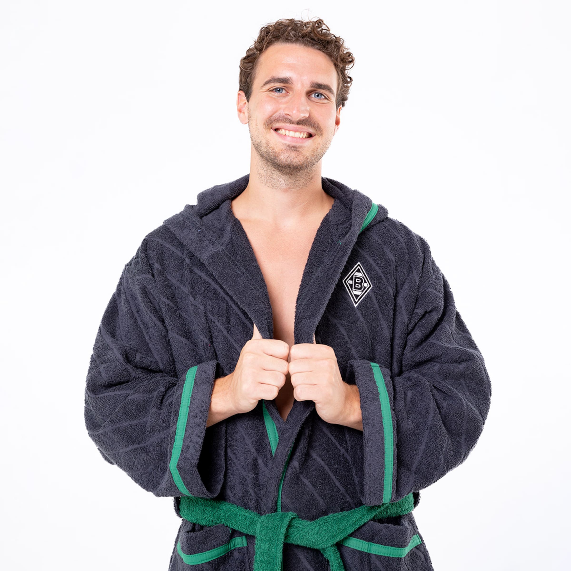 Ein Mann in einem dunkelgrauen weichen Bademantel mit grünem Gürtel und einem Borussia Mönchengladbach-Logo lächelt in die Kamera.