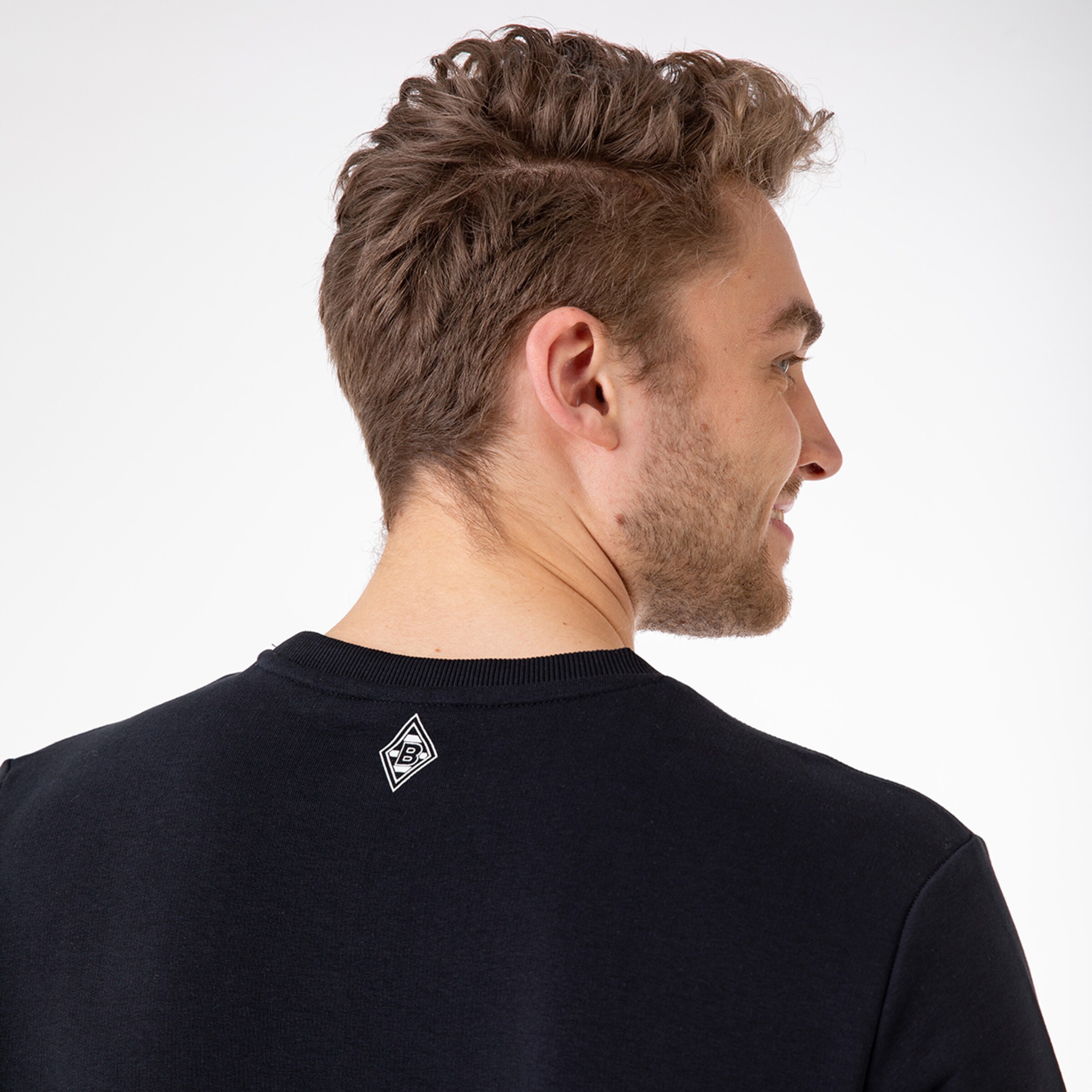 Vorderansicht eines Mannes mit lockigem Haar der lächelt und sich seitlich dreht in einem schwarzen Sweatshirt mit Vereinslogo.