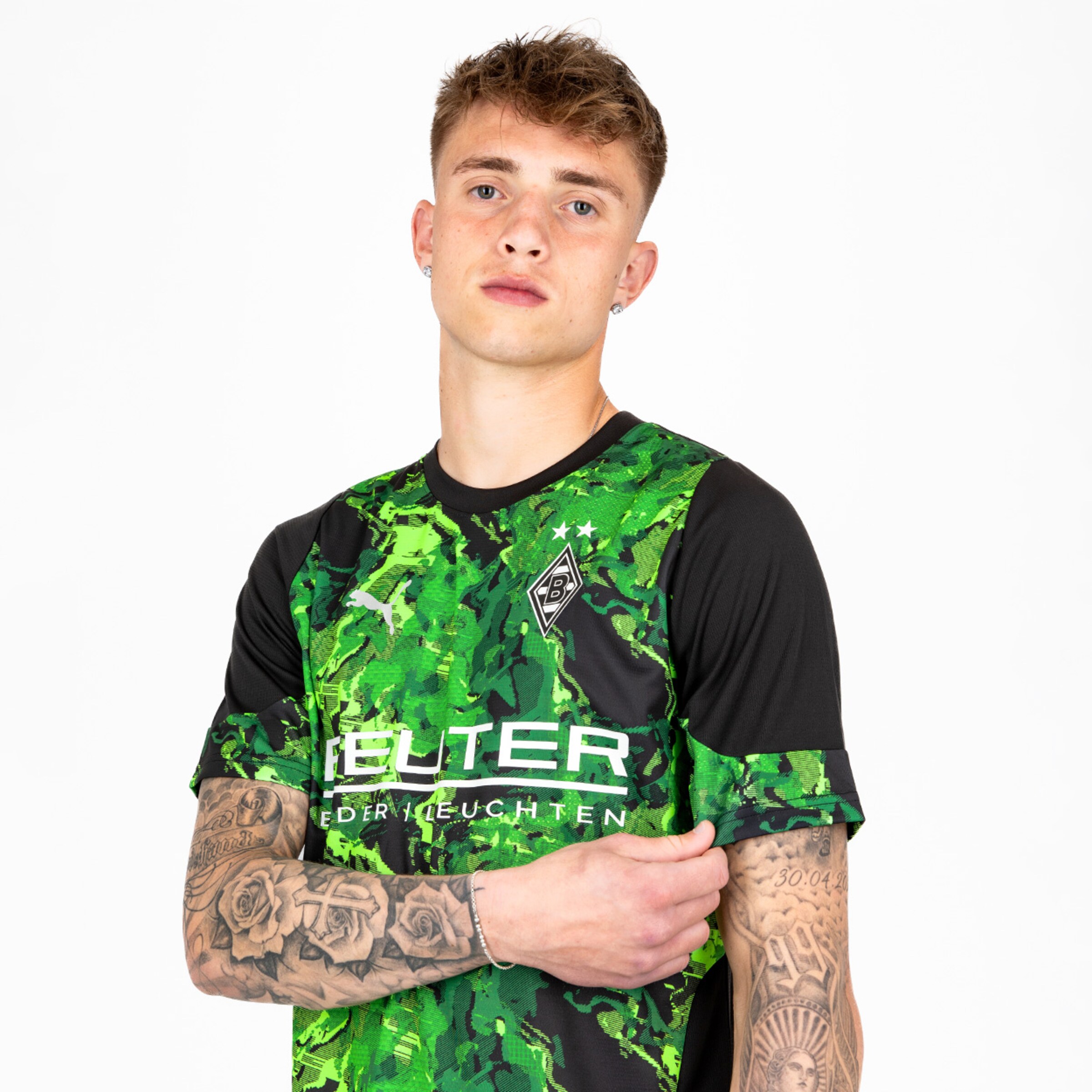 Junger Mann mit Tattoos in einem grünen Borussia Mönchengladbach Trikot stellt seine Arme verschränkt.