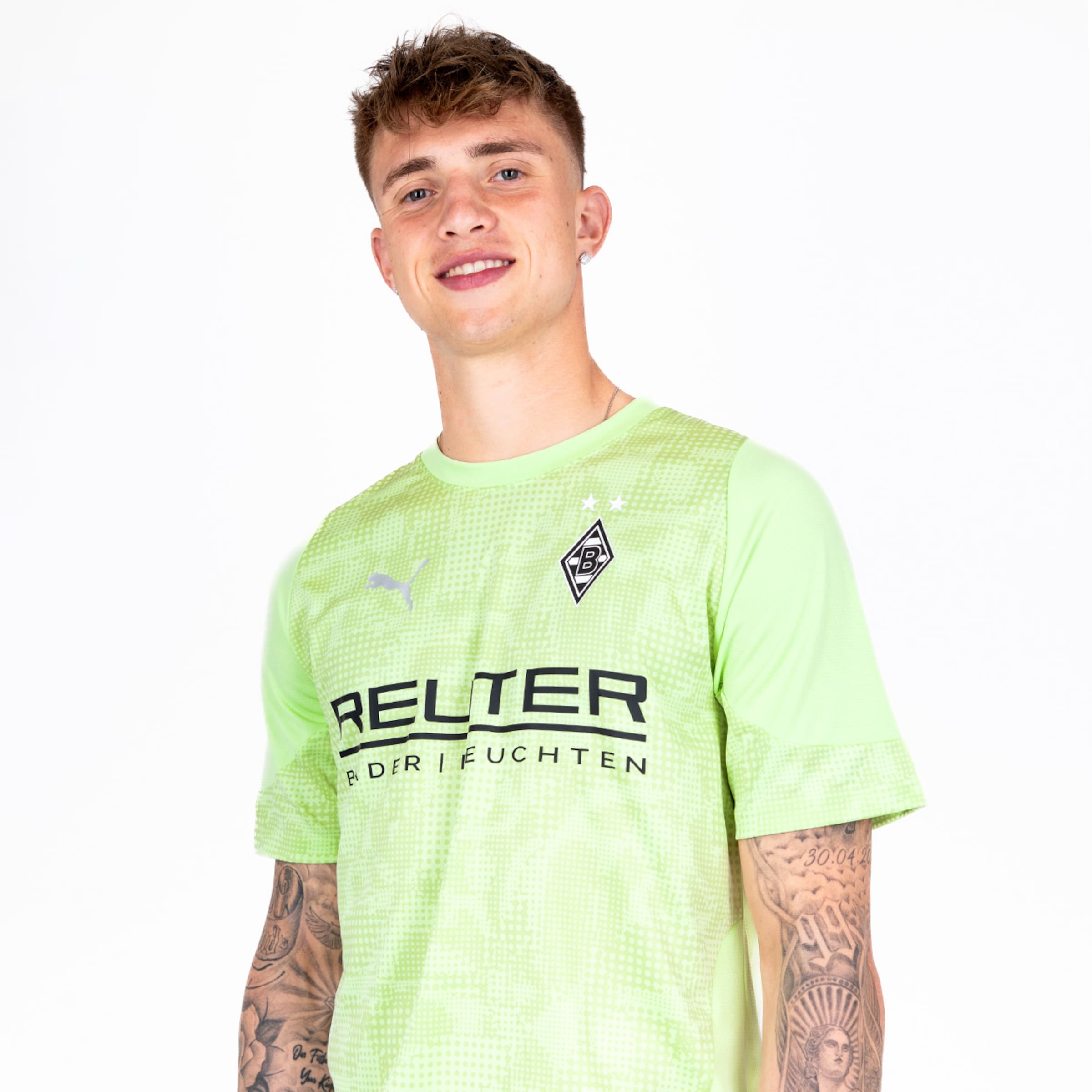 Ein Spieler in grünem Trikot mit aufgedrucktem Sponsorennamen steht lächelnd vor neutralem Hintergrund.