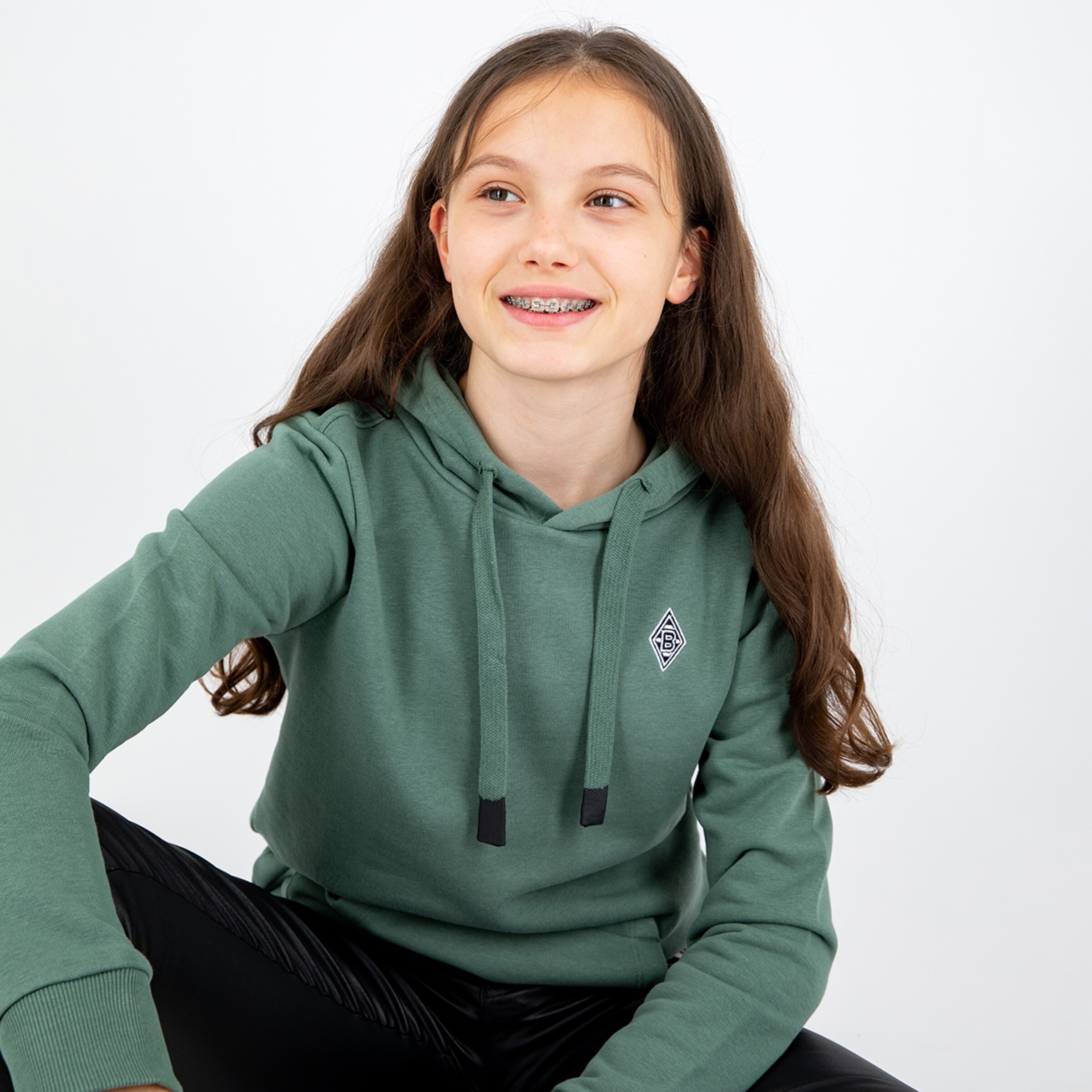 Mädchen mit langen braunen Haaren trägt einen grünen Hoodie mit Kapuze und lächelt.