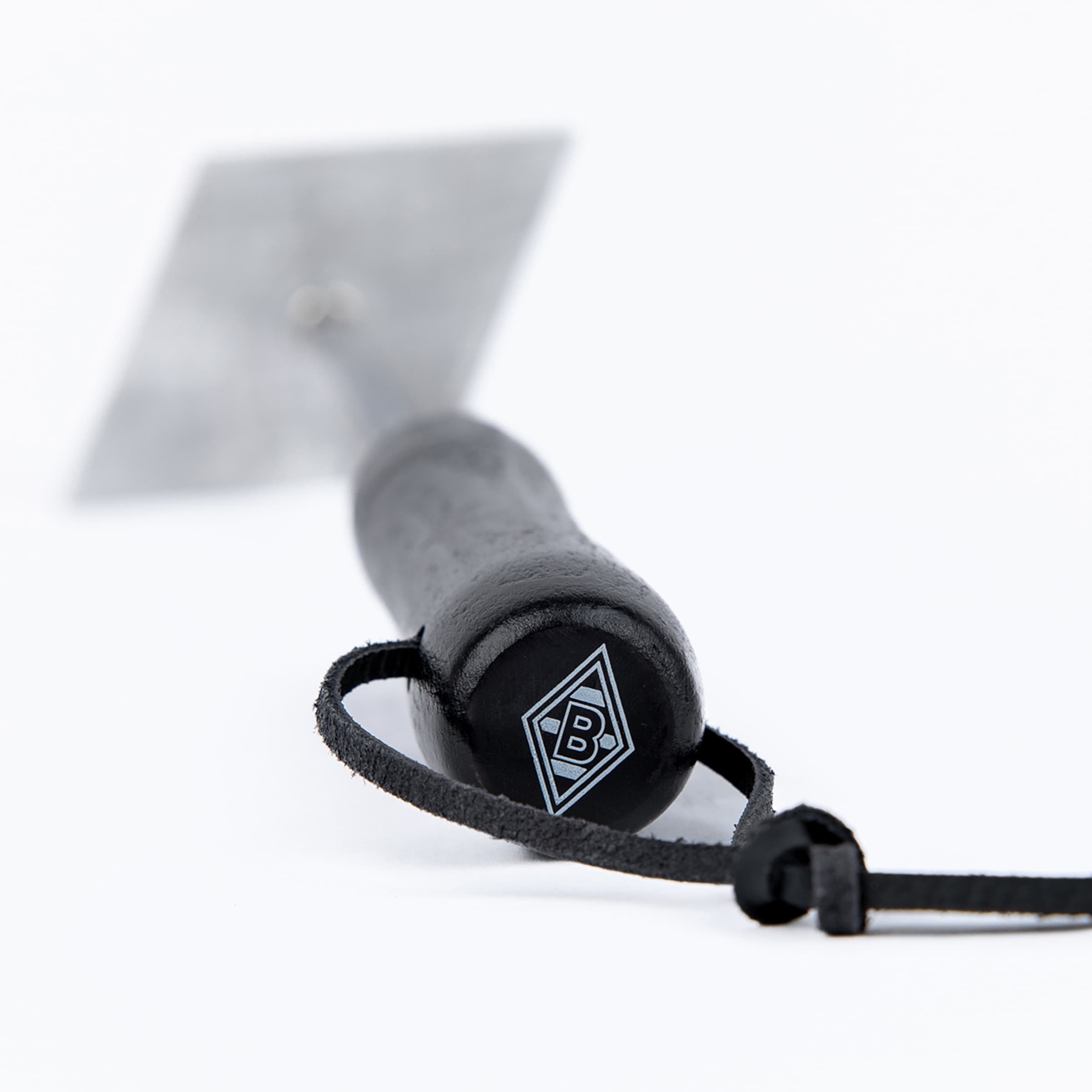 Nahaufnahme eines schwarzen Werkzeuggriffs mit B-Logo in einer Raute und Lederschlaufe. Unscharfe Metallklinge im Hintergrund auf weißem Grund.