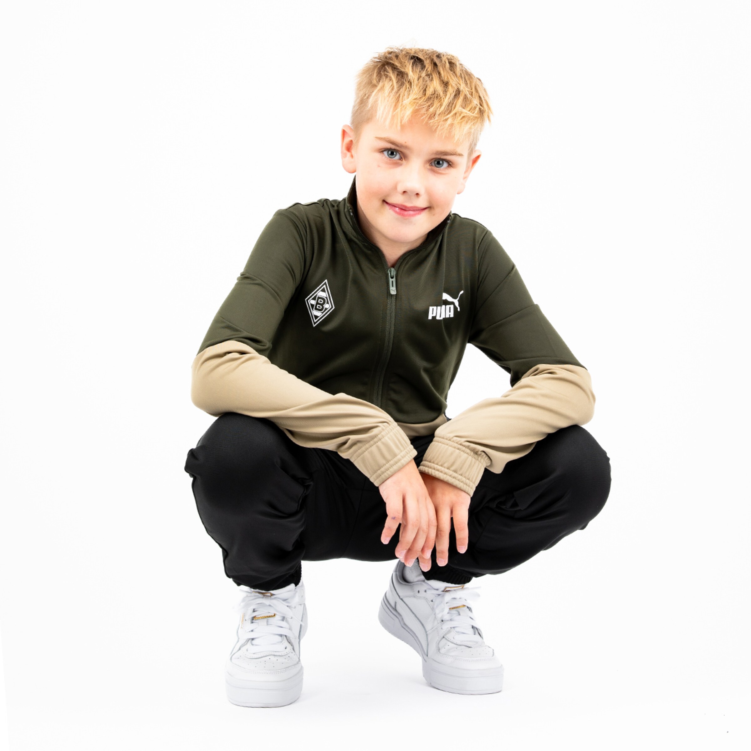 Ein Junge mit blonden Haaren in einem olivgrünen Sportoberteil und schwarzen Jogginghosen der sich hockend mit einem Lächeln präsentiert.