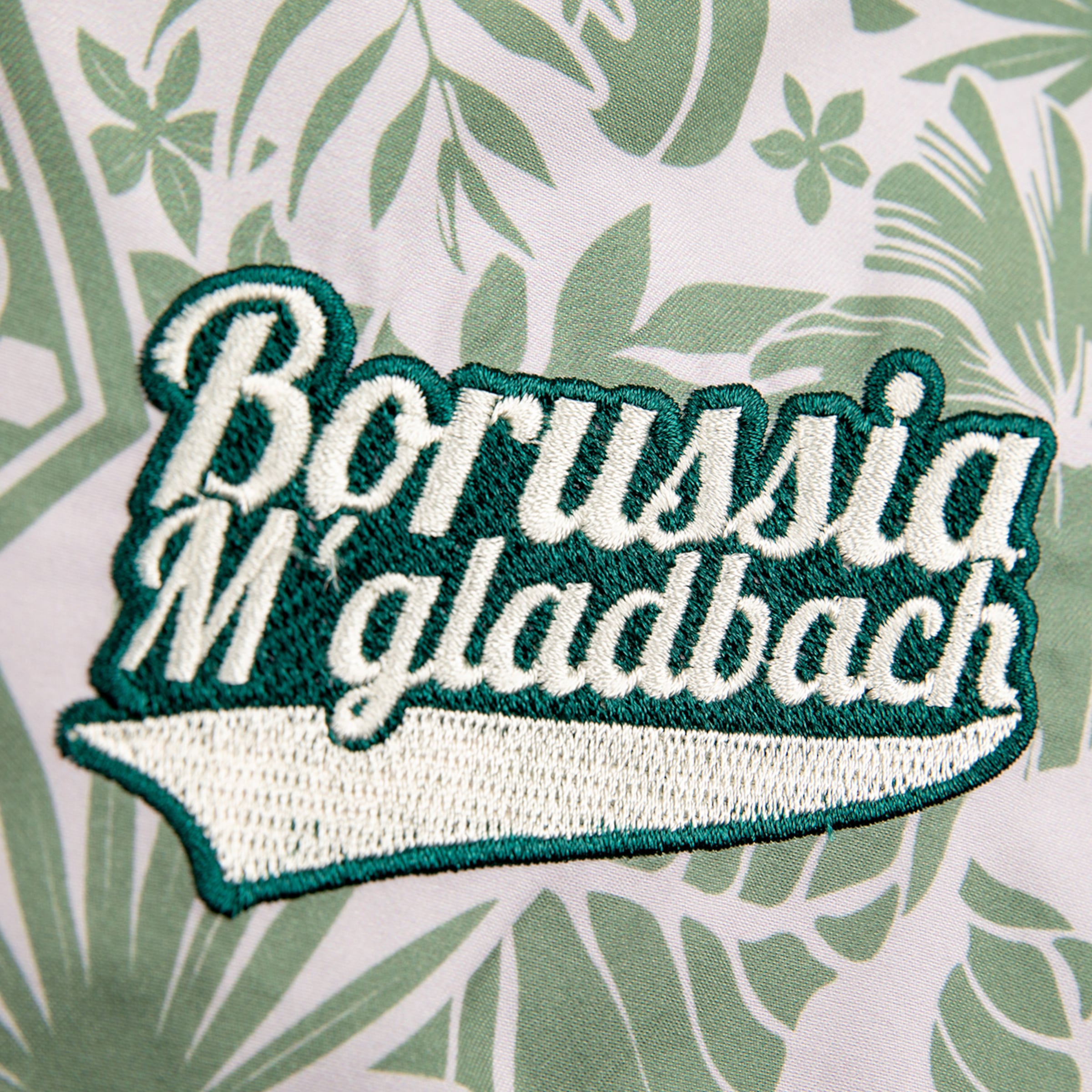 Nahaufnahme eines bestickten Borussia M'gladbach Logos in Weiß mit dunkelgrünem Rand, auf hellbeigem Stoff mit grünem Blattmuster im Hintergrund.