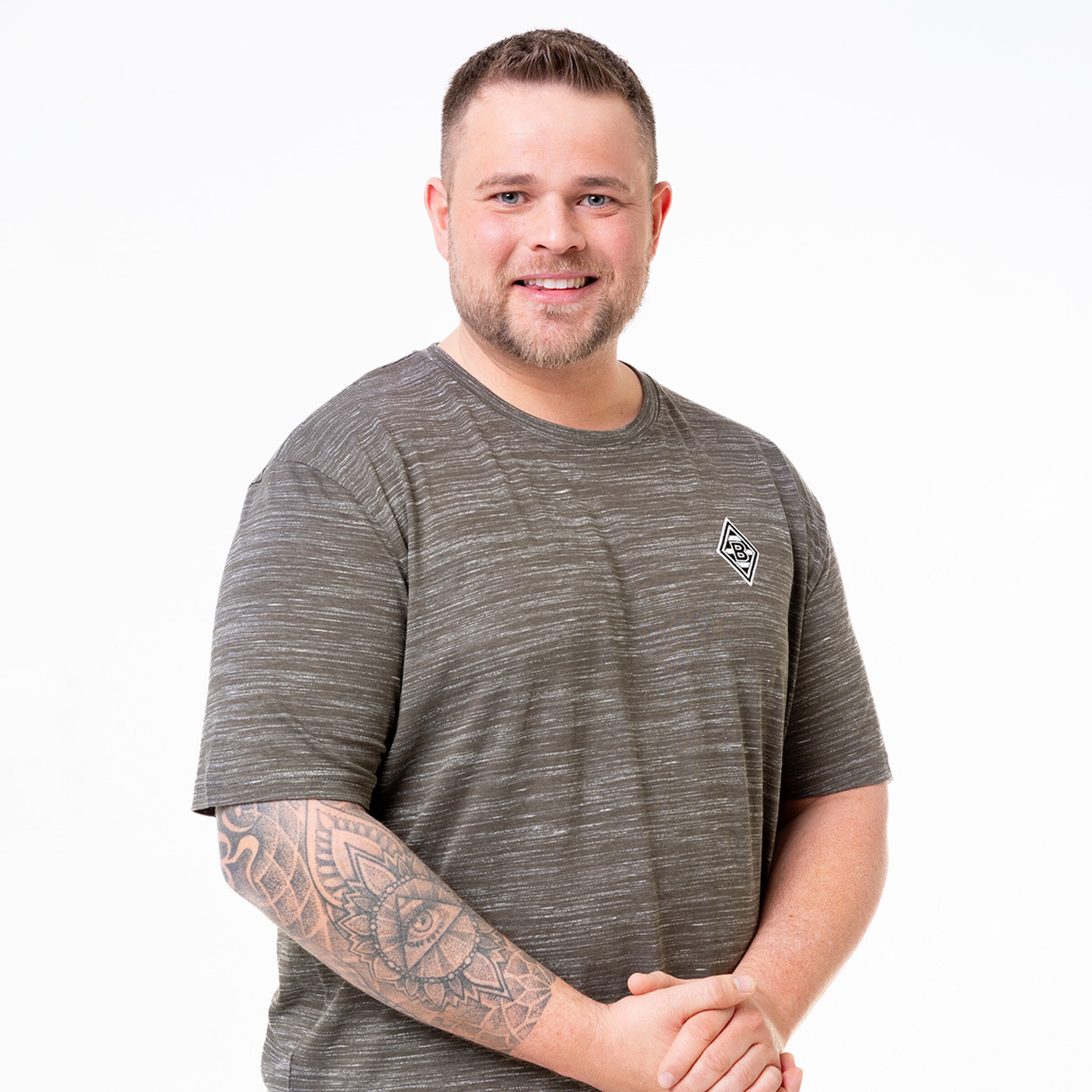 Mann mit tattoos grauem T-Shirt und Lächeln steht mit gefalteten Händen.