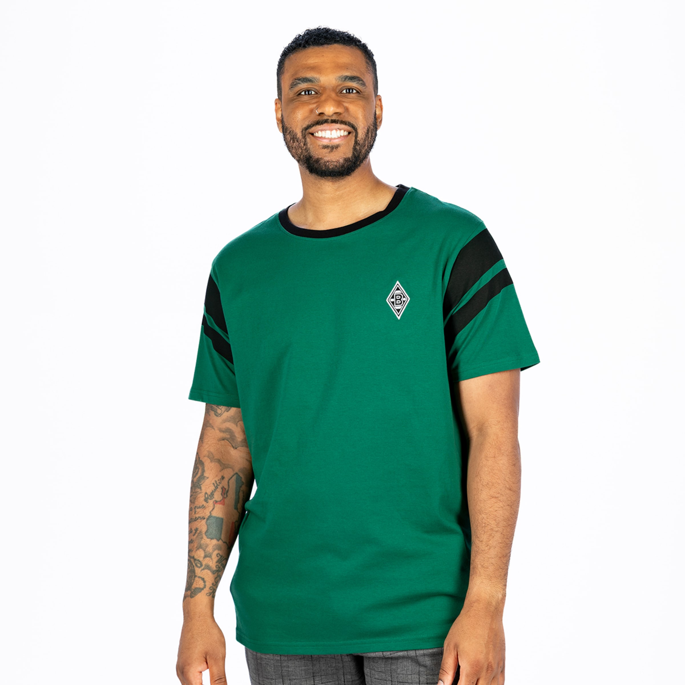 Lächelnder Mann mit Bart und Nasenpiercing, trägt grünes T-Shirt mit schwarzen Streifen an Ärmeln und Rautenlogo, dazu graue Karohose, vor weißem Hintergrund.