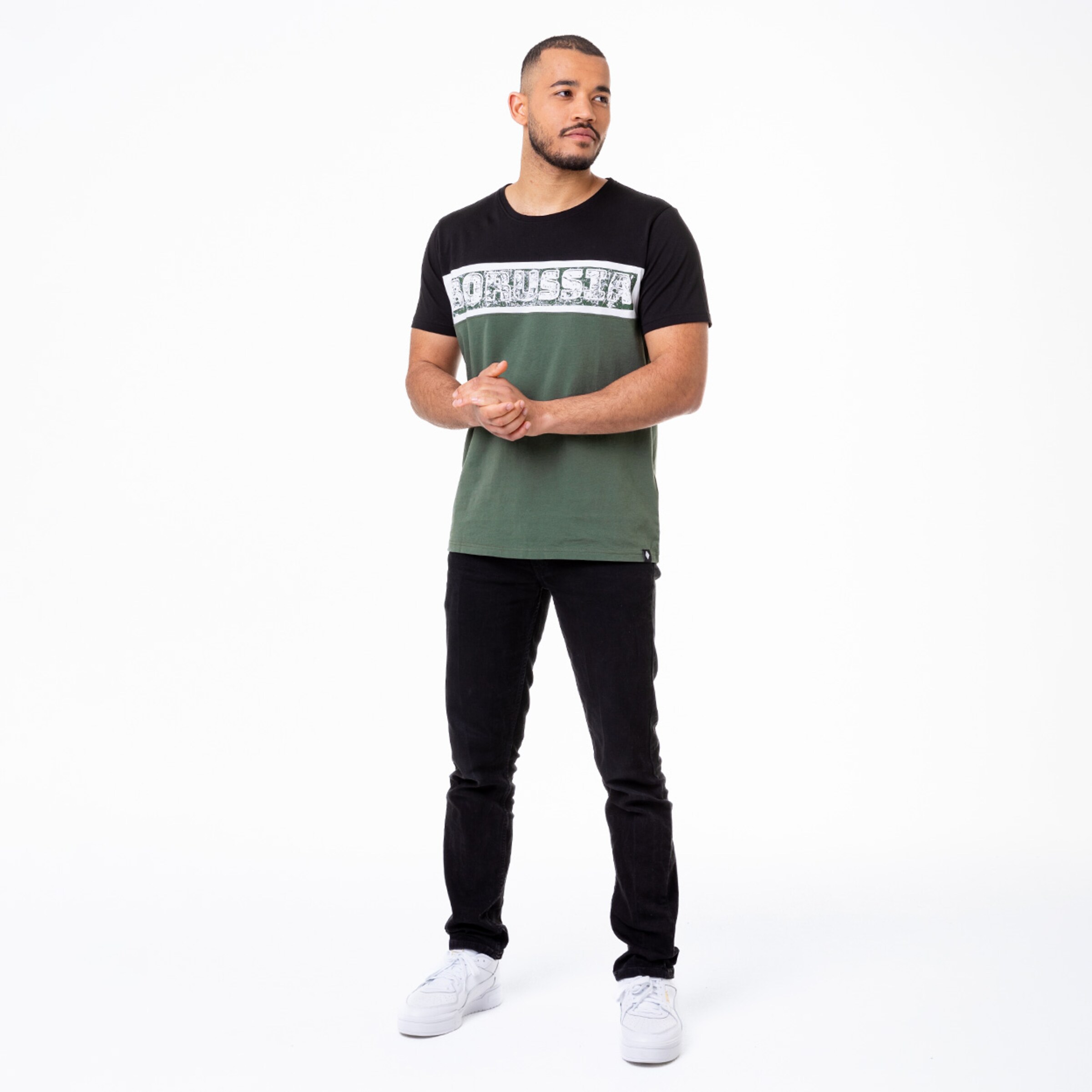 Mann in T-Shirt mit grün-schwarzem Design und schwarzer Hose steht mit verschränkten Händen.