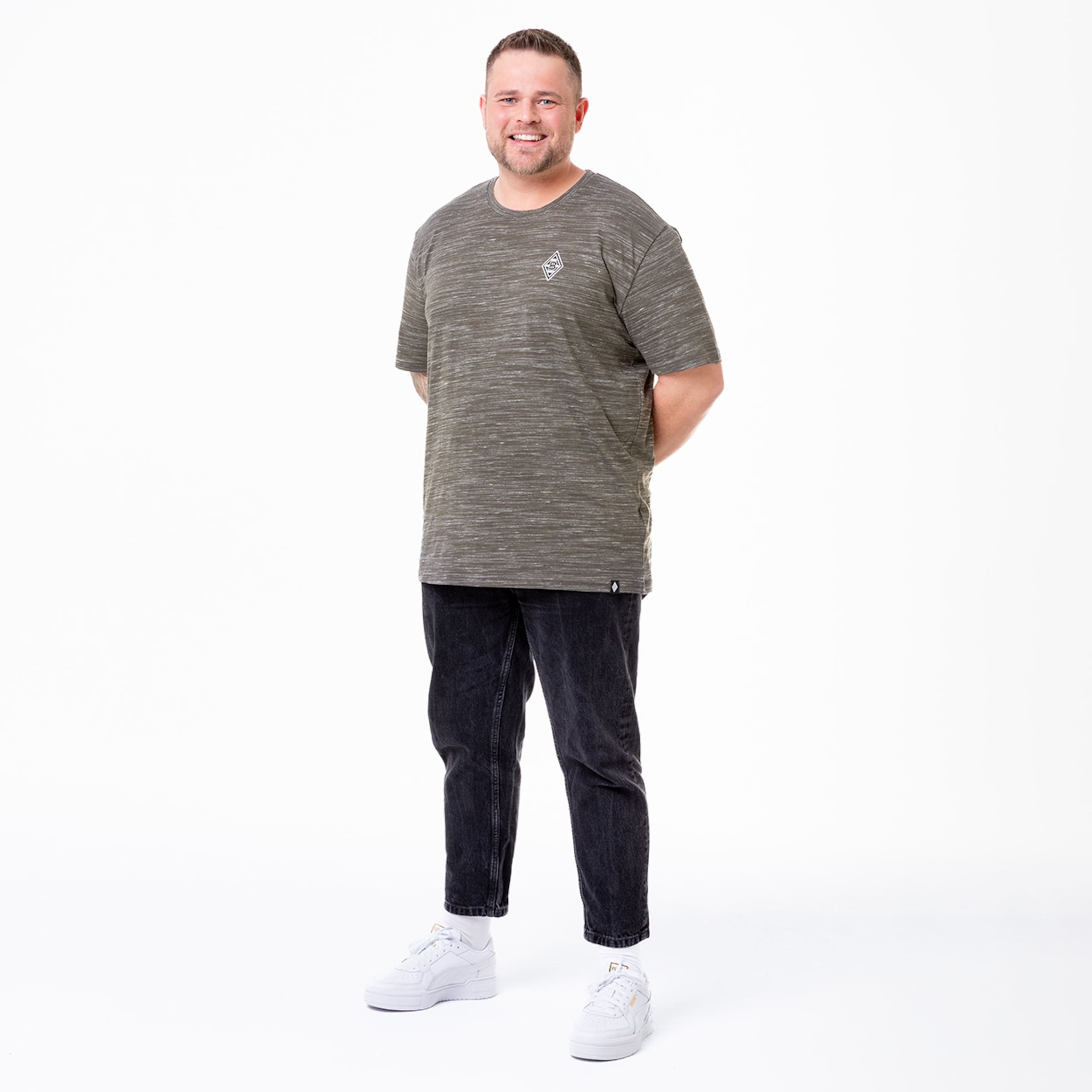 Ein Mann steht lächelnd mit einer Hand hinter dem Rücken. Er trägt ein graues gestreiftes T-Shirt schwarze Jeans und weiße Sneakers.