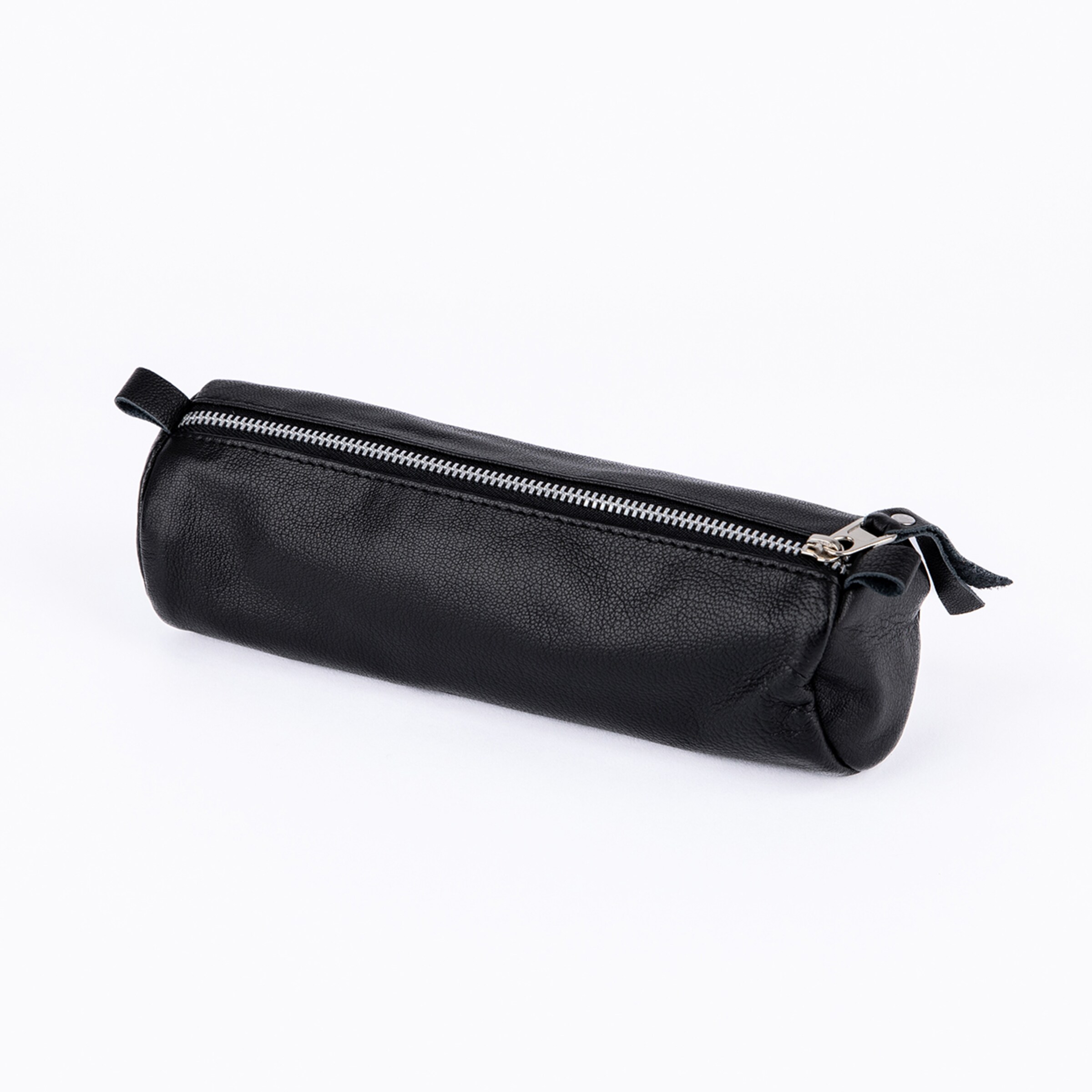 A black cylindrical pencil case made of pebbled leather is lying on a white background. It features a silver metal zipper along the top with a leather pull tab and a small leather loop at one end.