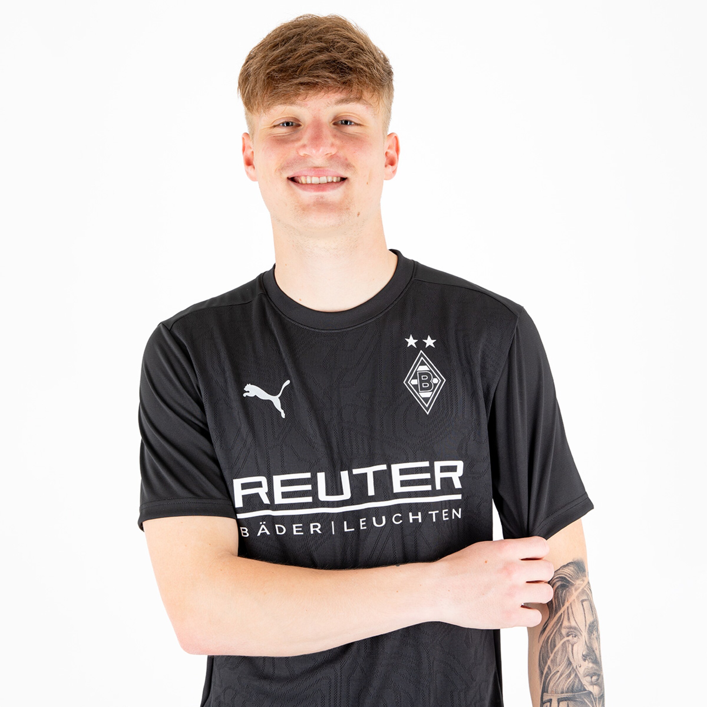 Junger Fußballspieler mit einem breiten Lächeln trägt das schwarze Heimtrikot von Borussia Mönchengladbach mit Sponsorennamen.