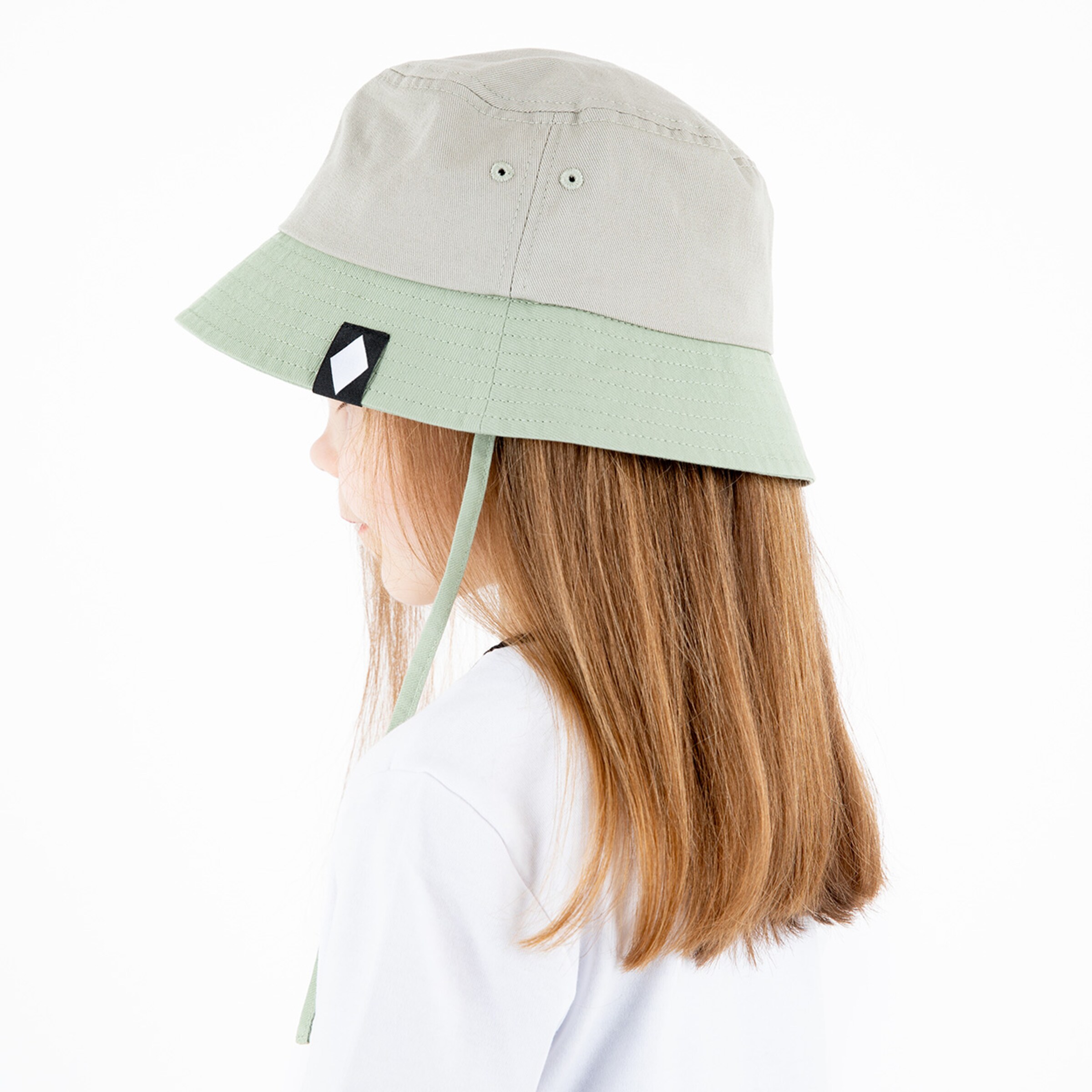 Mädchen mit braunen Haaren trägt einen beigen Hut mit grünem Rand zur Seite der eine Schnur hat.