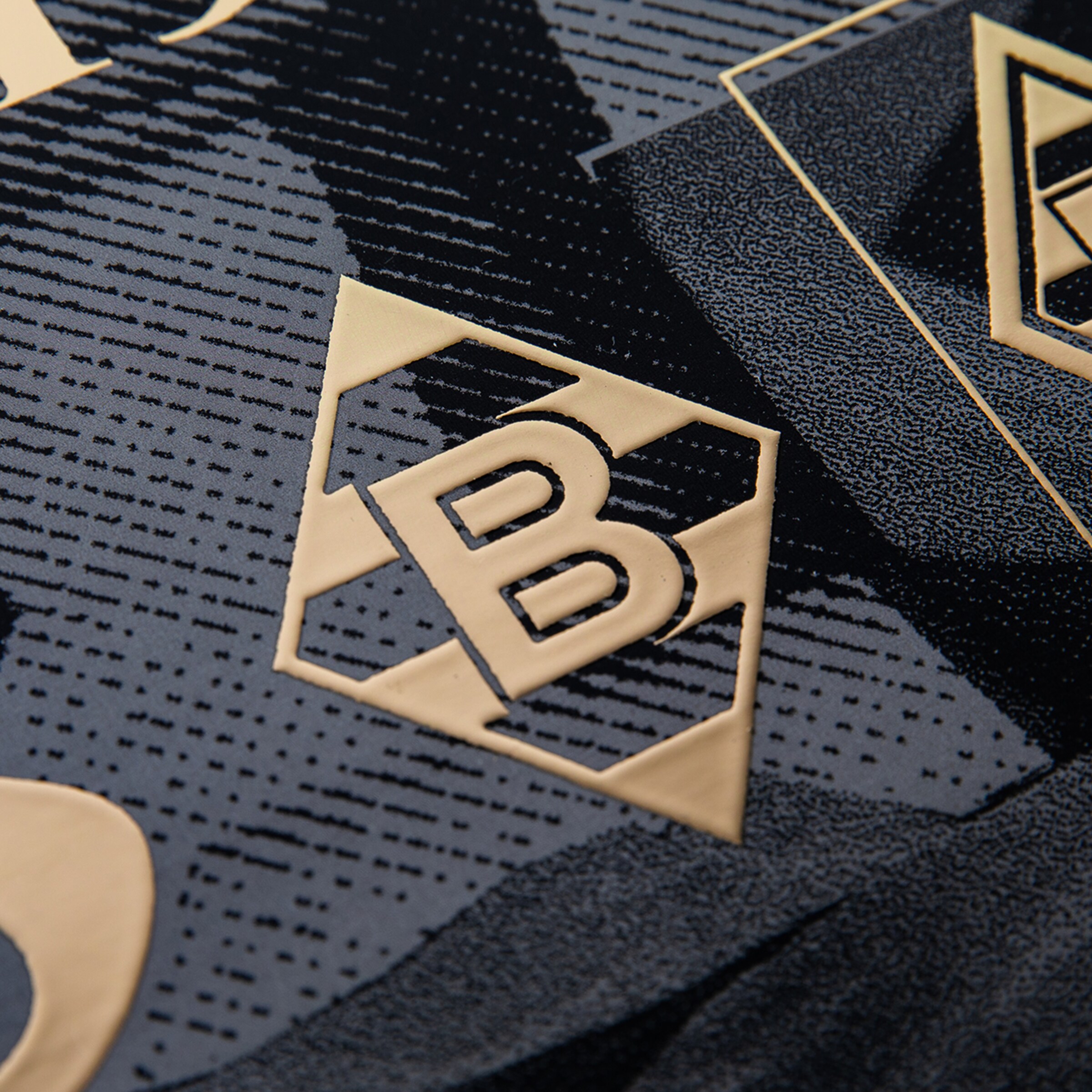 Nahaufnahme eines geprägten, goldfarbenen B-Logos in einer Raute auf dunkel gemustertem, strukturiertem Stoff.