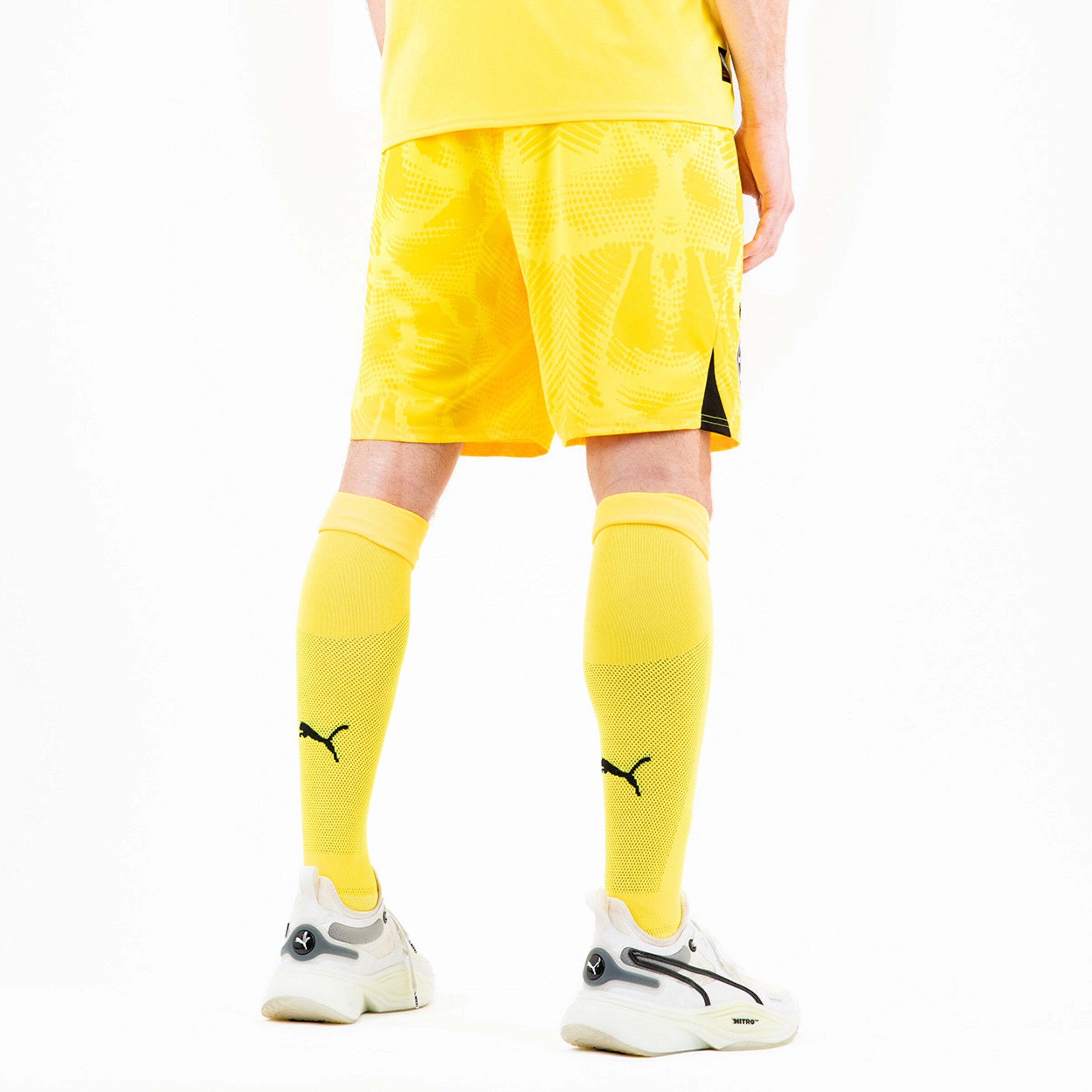 Ein Spieler trägt ein gelbes Trikot mit passenden kurzen Hosen und Socken während er auf den Füßen weiße Sportchuhe mit schwarzem Logo steht.