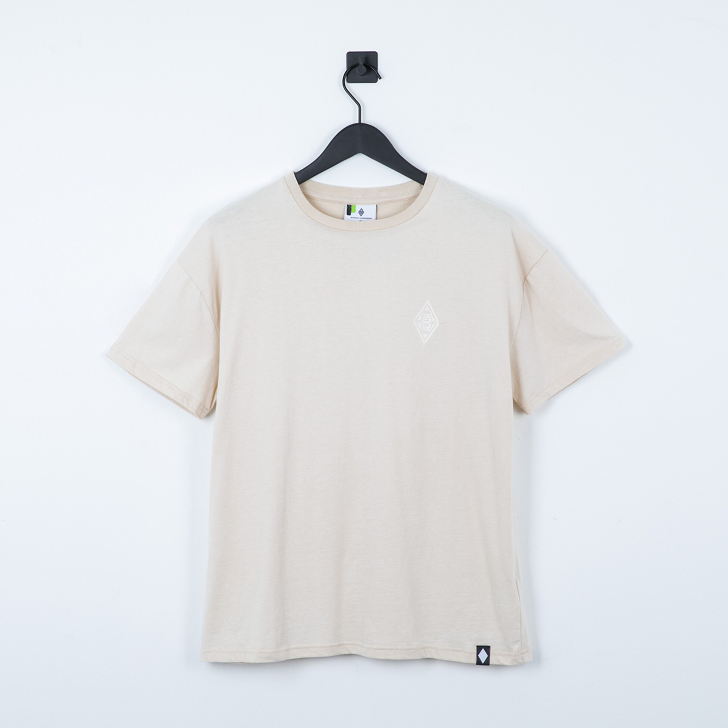 Beiges T-Shirt mit weißem Rauten-Logo auf der Brust und Label am Saum, hängend an einem schwarzen Kleiderbügel vor weißem Hintergrund.