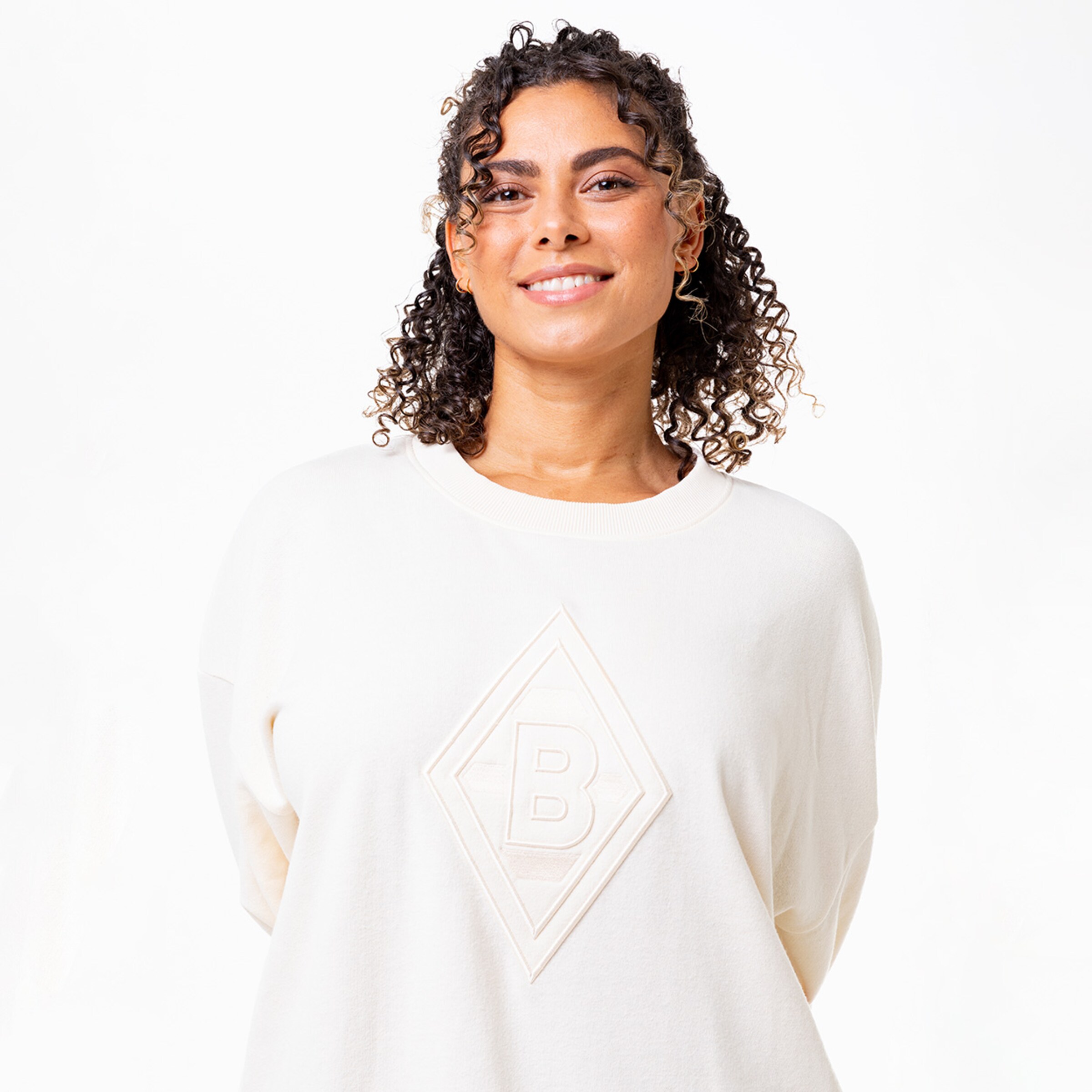 Frau mit lockigem Haar steht lächelnd in einem hellen Sweatshirt mit dem Logo der Borussia Mönchengladbach.