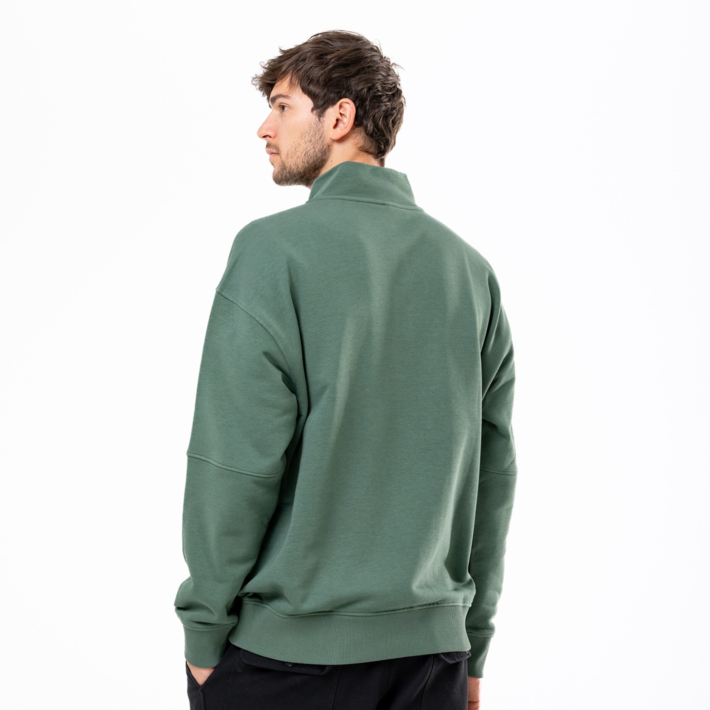 Rückansicht eines Mannes in grünem Half-Zip-Sweatshirt mit überschnittenen Schultern und schwarzen Hosen vor weißem Hintergrund.