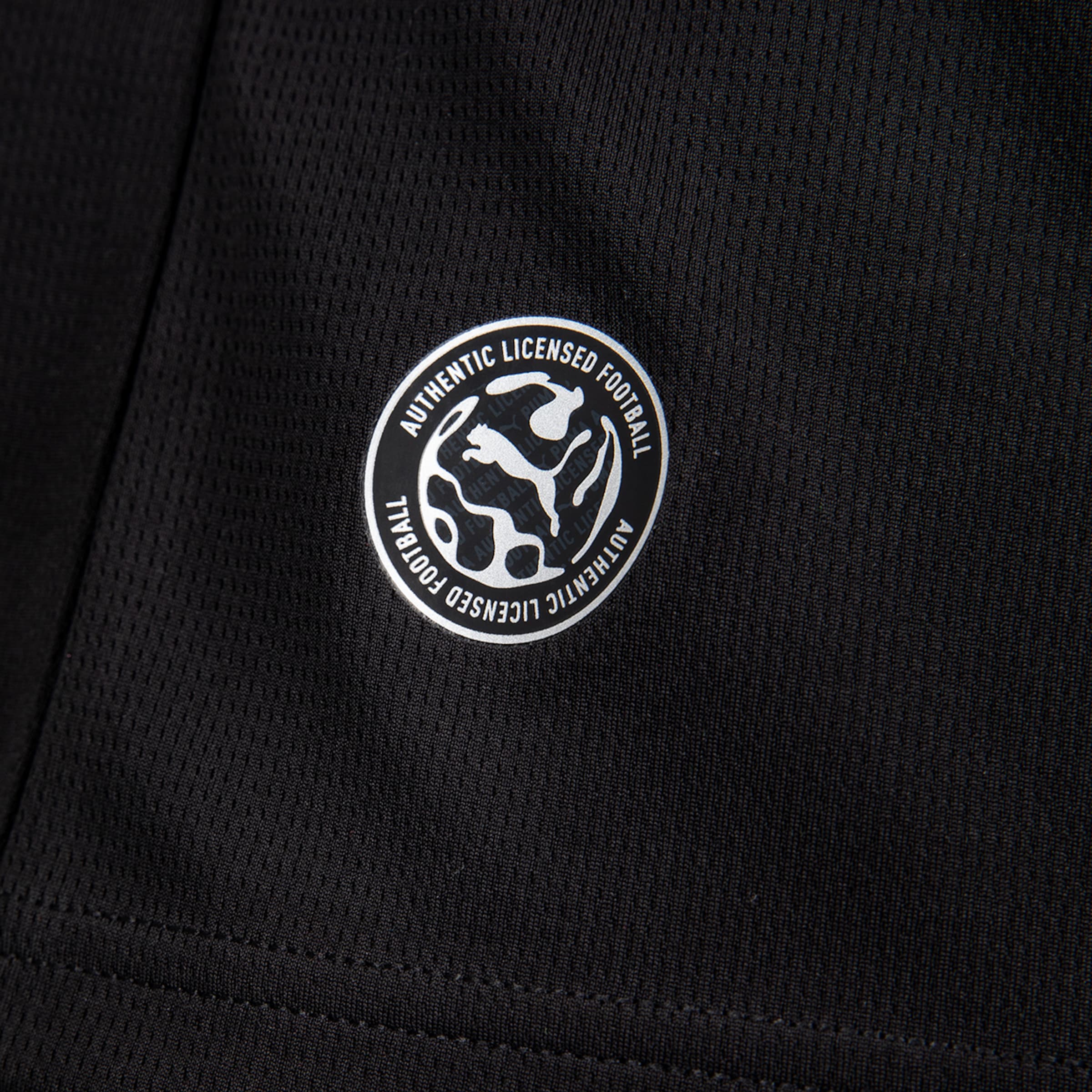 Nahaufnahme von schwarzem Mesh-Gewebe mit rundem silbernem Emblem, das ein Puma-Logo und den Text „AUTHENTIC LICENSED FOOTBALL“ zeigt.