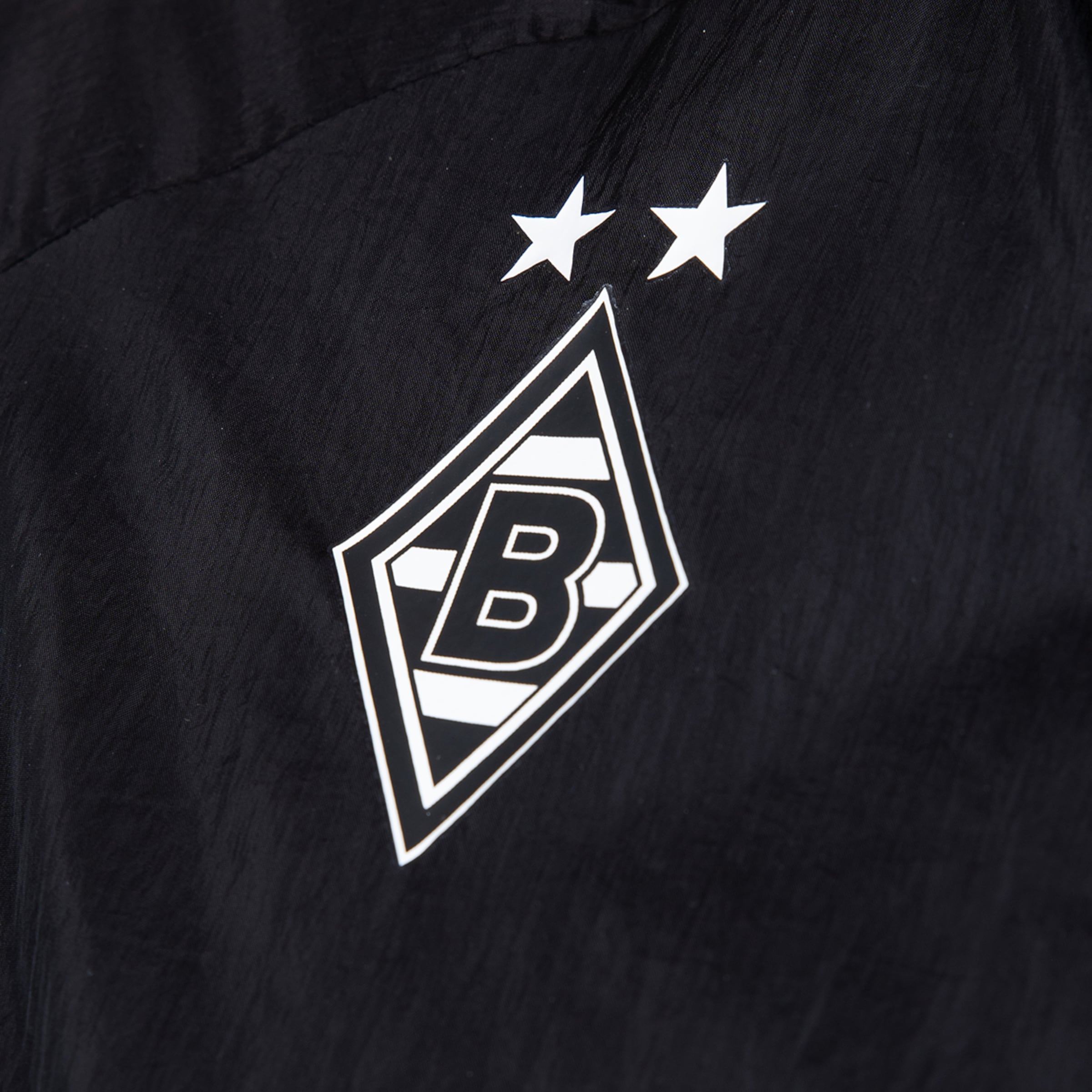 Logo von Borussia Mönchengladbach: Eine weiße Raute mit schwarzem 'B' und zwei Sternen auf schwarzem Textilgewebe.