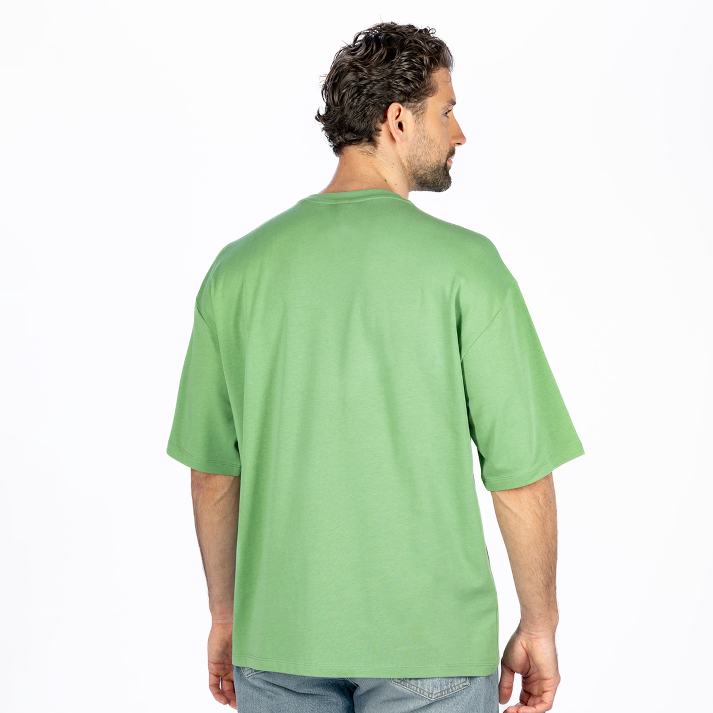 Rear view of a man with dark curly hair and a beard, wearing a plain green oversized t-shirt and blue jeans, turned slightly to his right against a white background.
