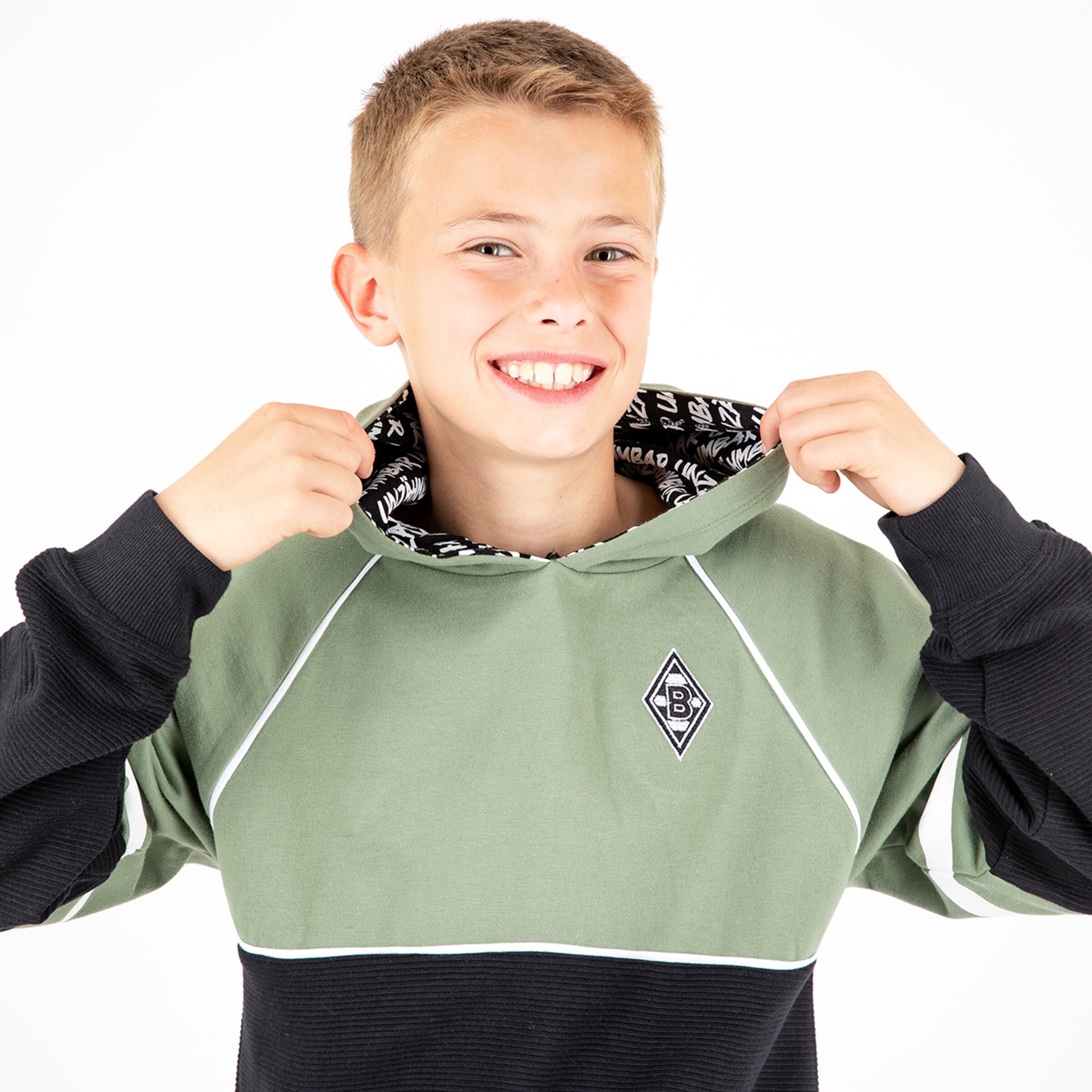 Junger Mann mit kurzem blondem Haar trägt einen Kapuzenpullover in Olivgrün und Schwarz. Er lächelt und zieht die Kapuze über seinen Kopf.