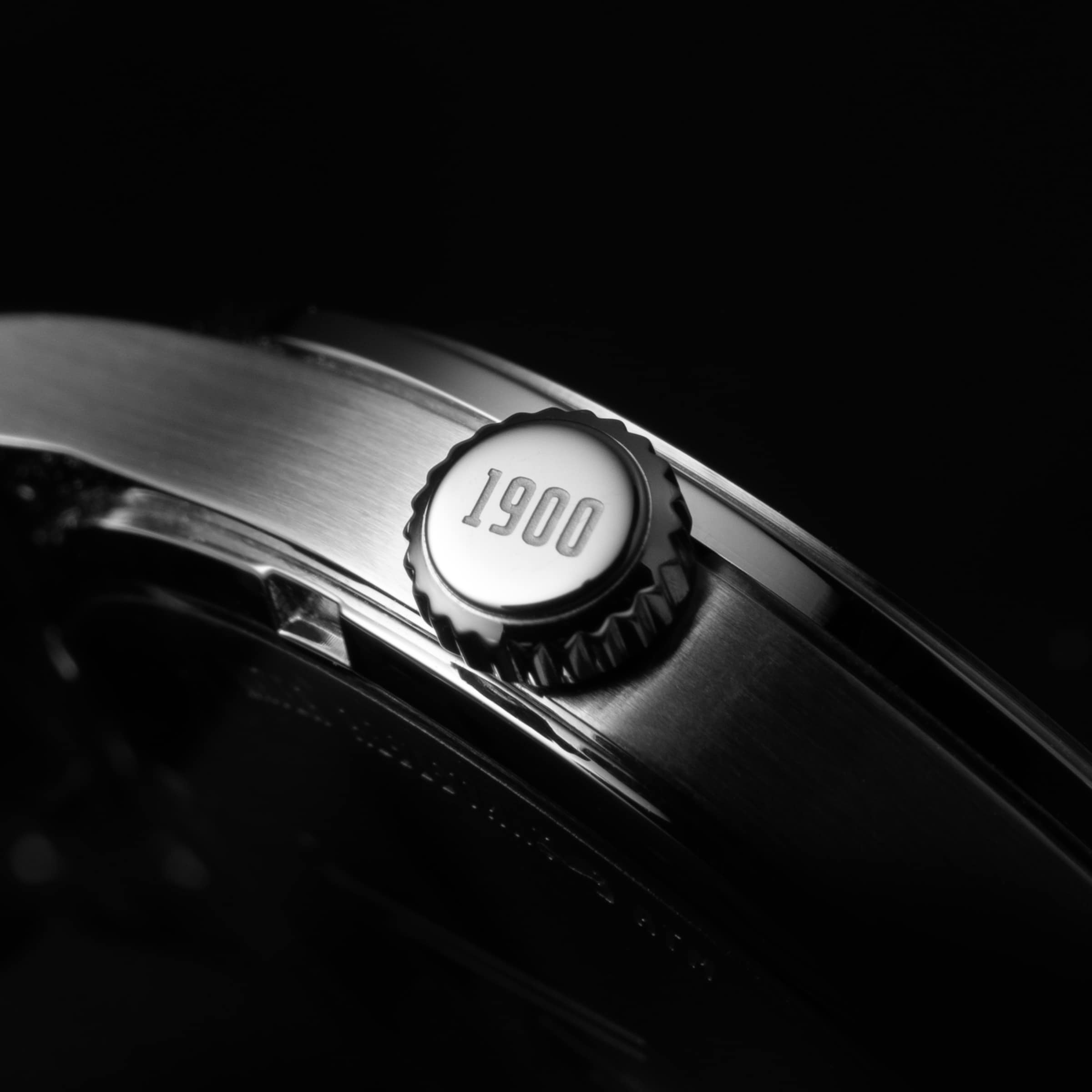 Nahaufnahme einer silbernen Uhrenkrone mit der Gravur „1900“. Schwarz-weiß-Foto mit elegantem Lichtspiel auf gebürstetem Metall.