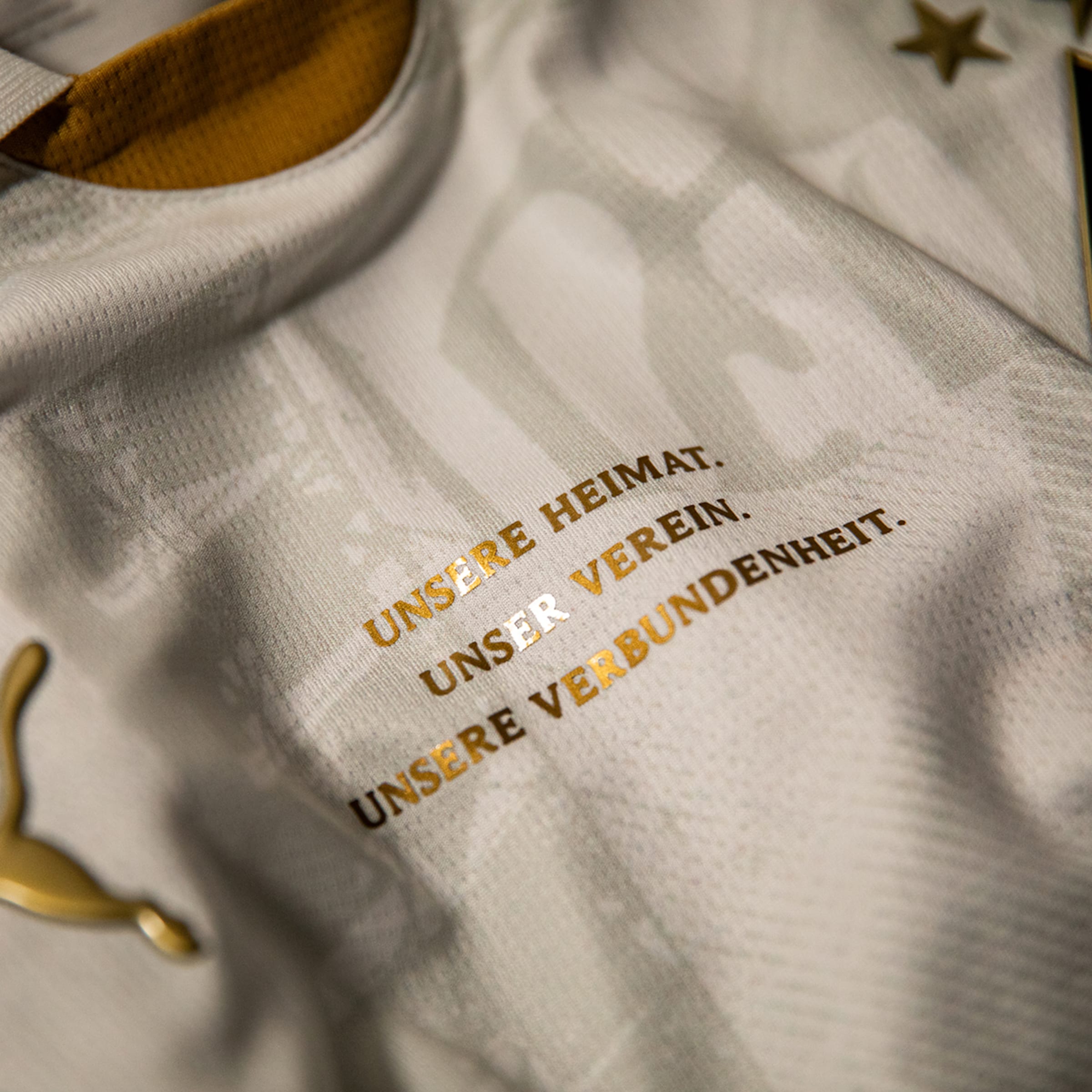 Nahaufnahme eines weißen Puma-Trikots mit dem goldenen Schriftzug „UNSERE HEIMAT. UNSER VEREIN. UNSERE VERBUNDENHEIT.“