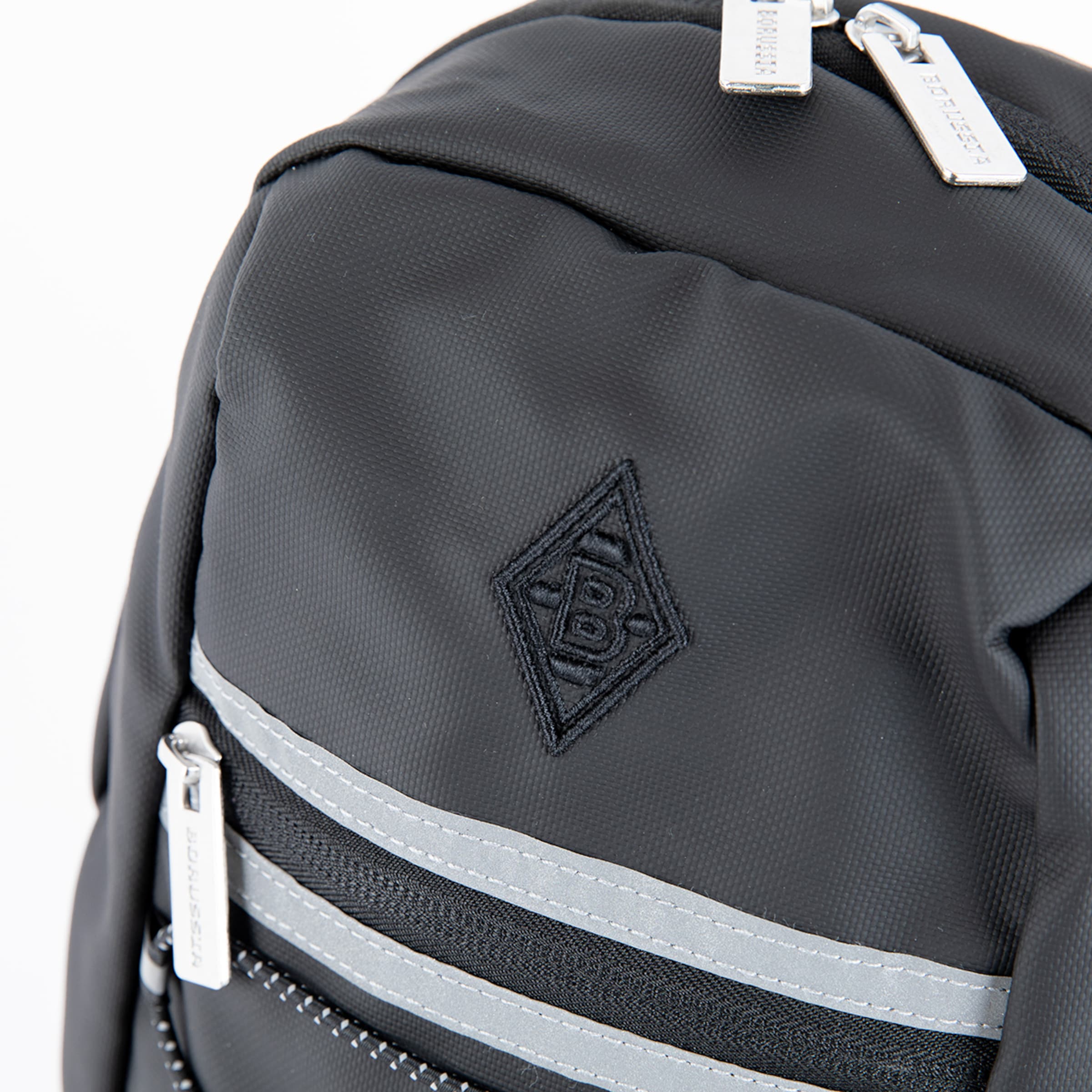 Detail eines schwarzen Rucksacks mit besticktem Borussia Mönchengladbach Rautenlogo, reflektierenden Streifen und silbernen Reißverschlüssen.