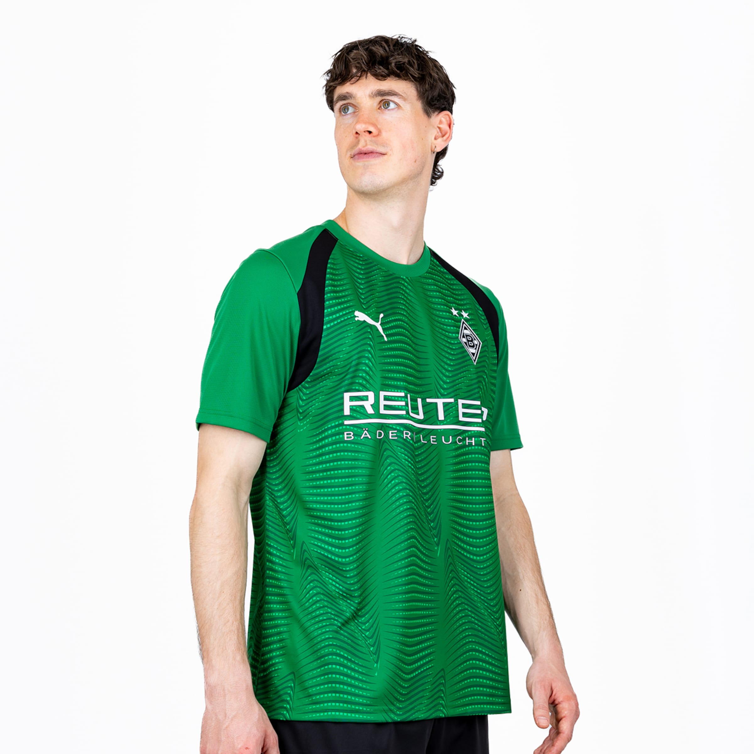 Ein Mann mit braunem Lockenhaar trägt ein grün-schwarzes Borussia Mönchengladbach Fußballtrikot mit welligem Muster, Puma-Logo, Vereinswappen mit zwei Sternen und dem Sponsor 'REUTE BÄDER LEUCHT'. Er blickt nach links vor weißem Hintergrund.