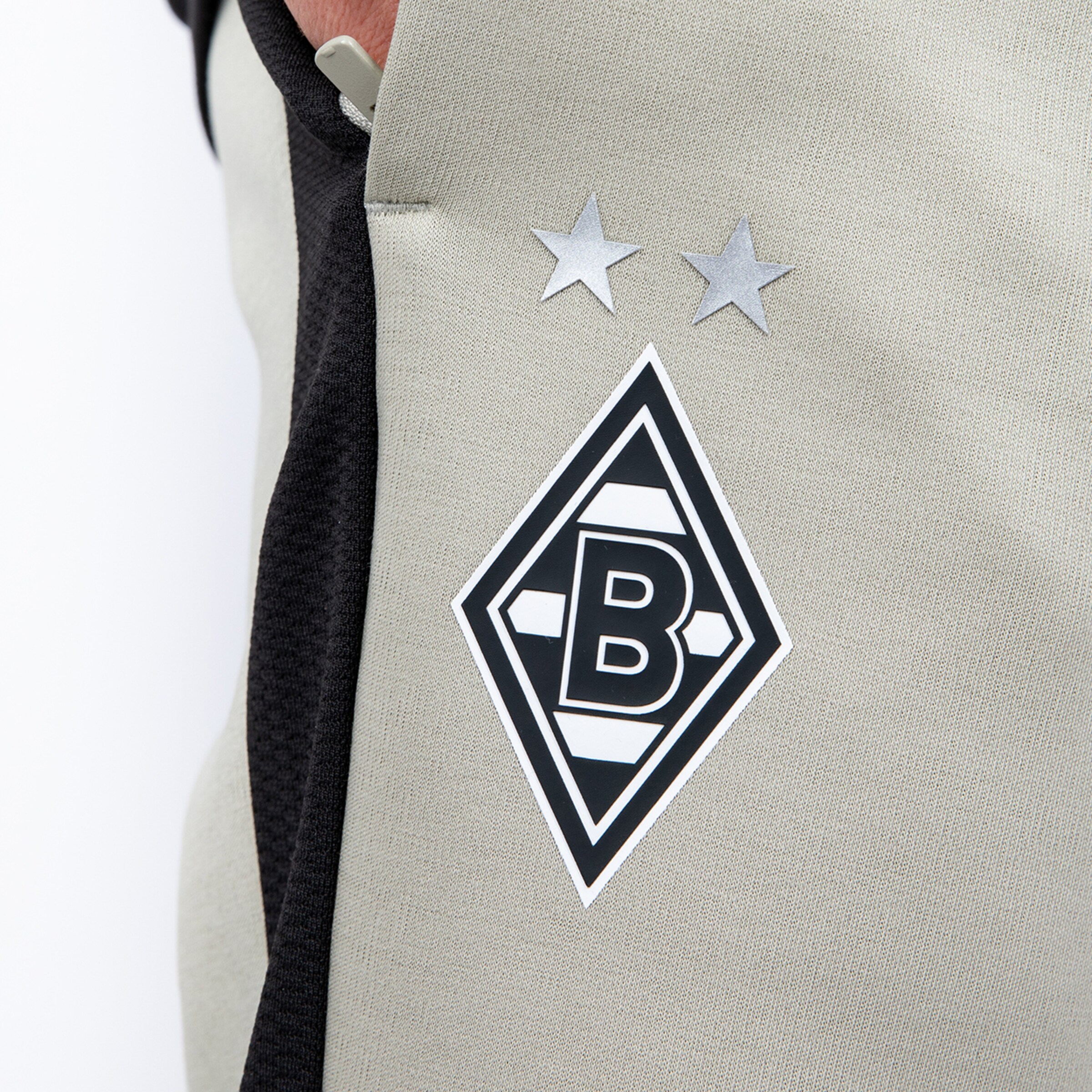 Nahaufnahme eines hellgrauen Stoffes mit einem schwarz-weißen rautenförmigen Logo mit dem Buchstaben 'B' und zwei silbernen Sternen darüber. Ein schwarzer Stoffstreifen ist links zu sehen.