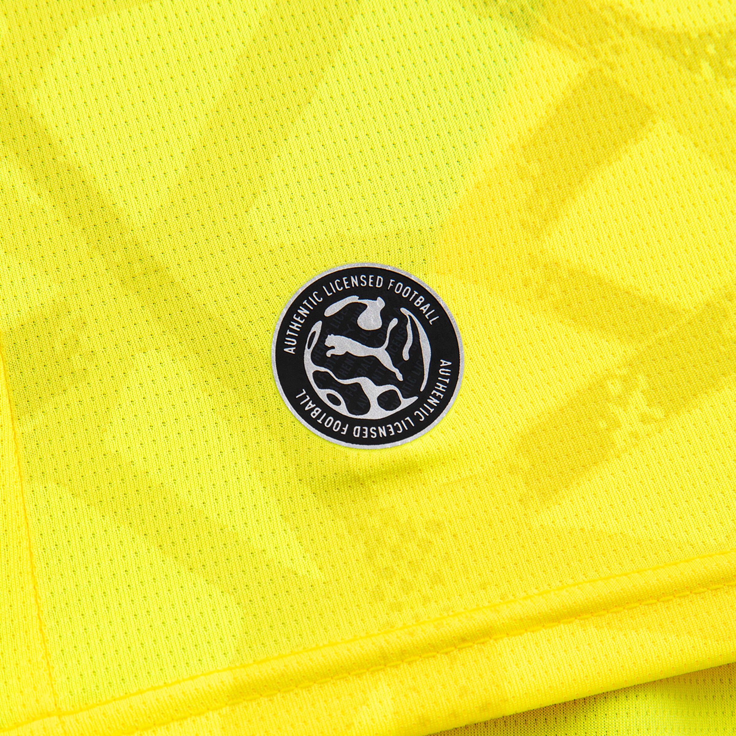 Nahaufnahme eines neongelben Trikots mit einem runden Puma-Patch „AUTHENTIC LICENSED FOOTBALL“.