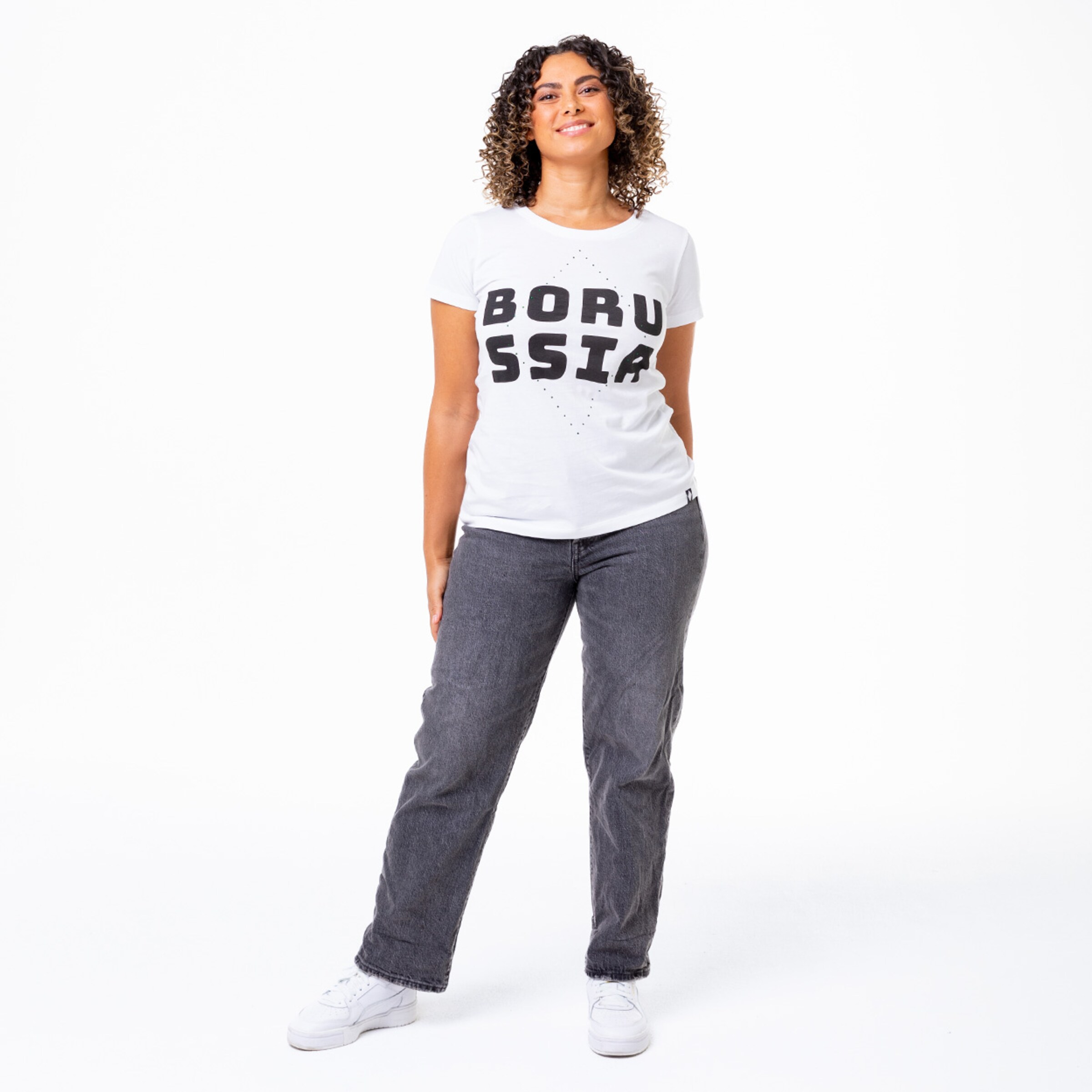 Woman stands wearing a white t-shirt with BORUSSIA lettering and gray jeans.